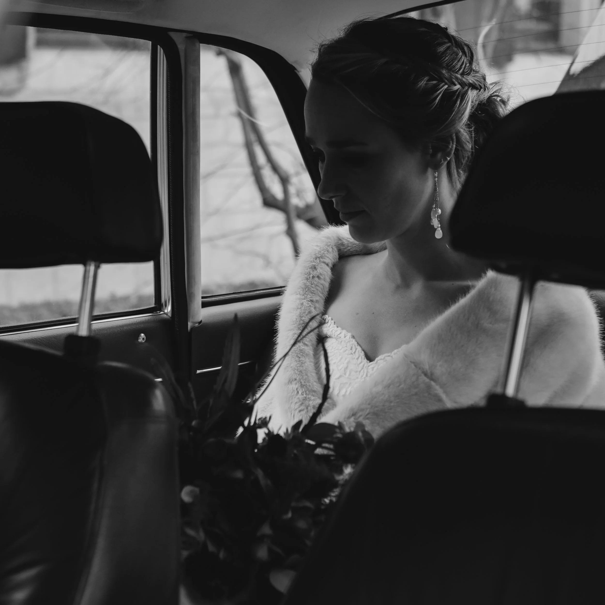 A woman with styled hair and dangling earrings sitting in the back seat of a car, looking downwards, with some flowers in the foreground.