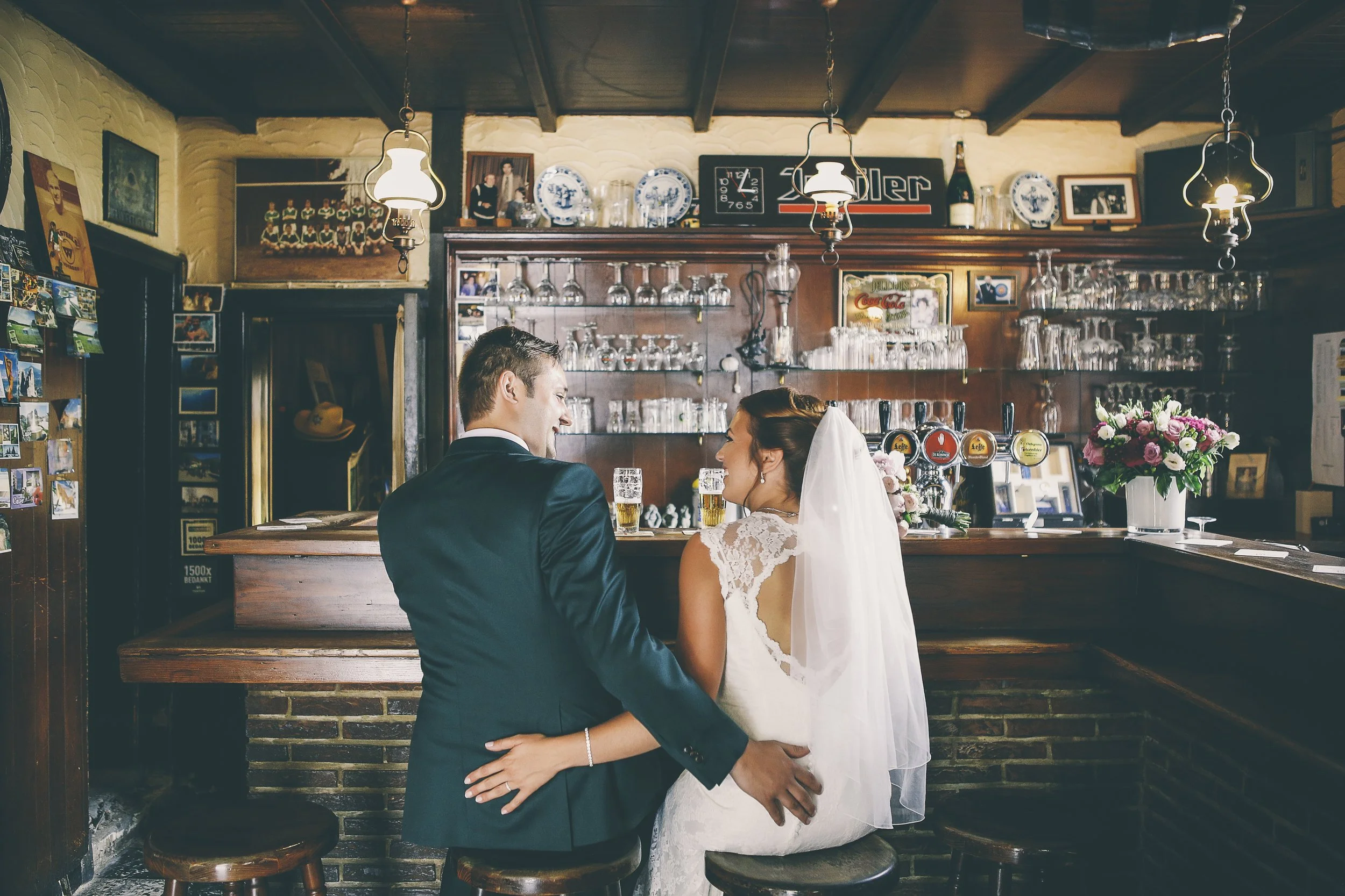 A bride and groom sitting together at a bar, smiling at each other. The bride is in a white wedding dress with a veil, and the groom is in a dark suit. There are drinks on the bar and various glassware and decorative items behind them.