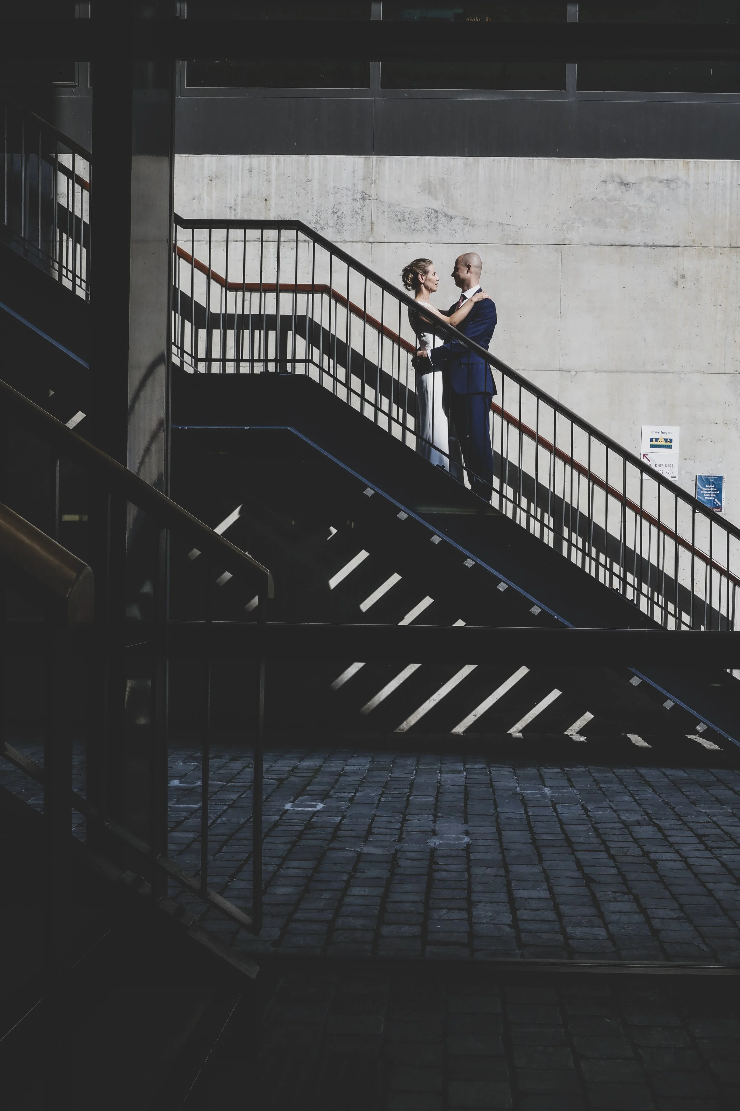 A bride and groom standing on an outdoor staircase, holding each other and looking into each other's eyes.