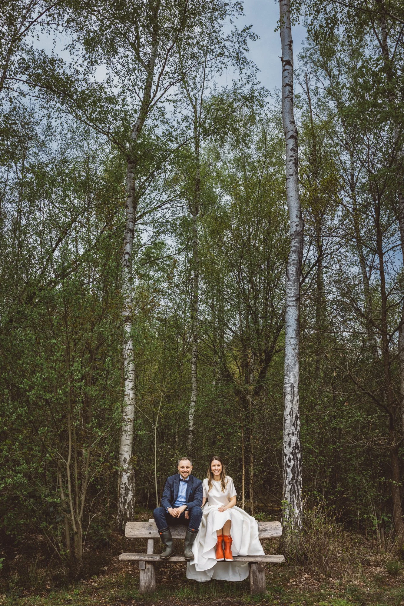 A couple sitting on a wooden bench in a forest with tall, thin trees and green leaves, smiling at the camera. The woman is wearing a white dress and orange rain boots, and the man is dressed in a dark suit with rain boots.