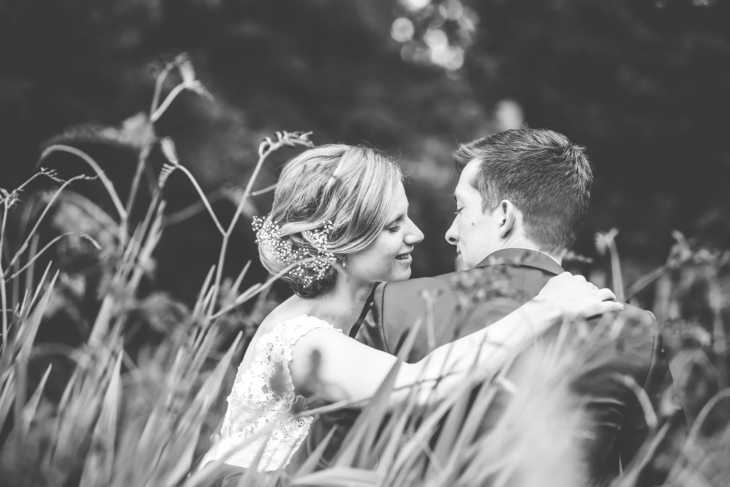 Black and white photo of a woman and a man embracing outdoors, facing each other with eyes closed, surrounded by tall grass.