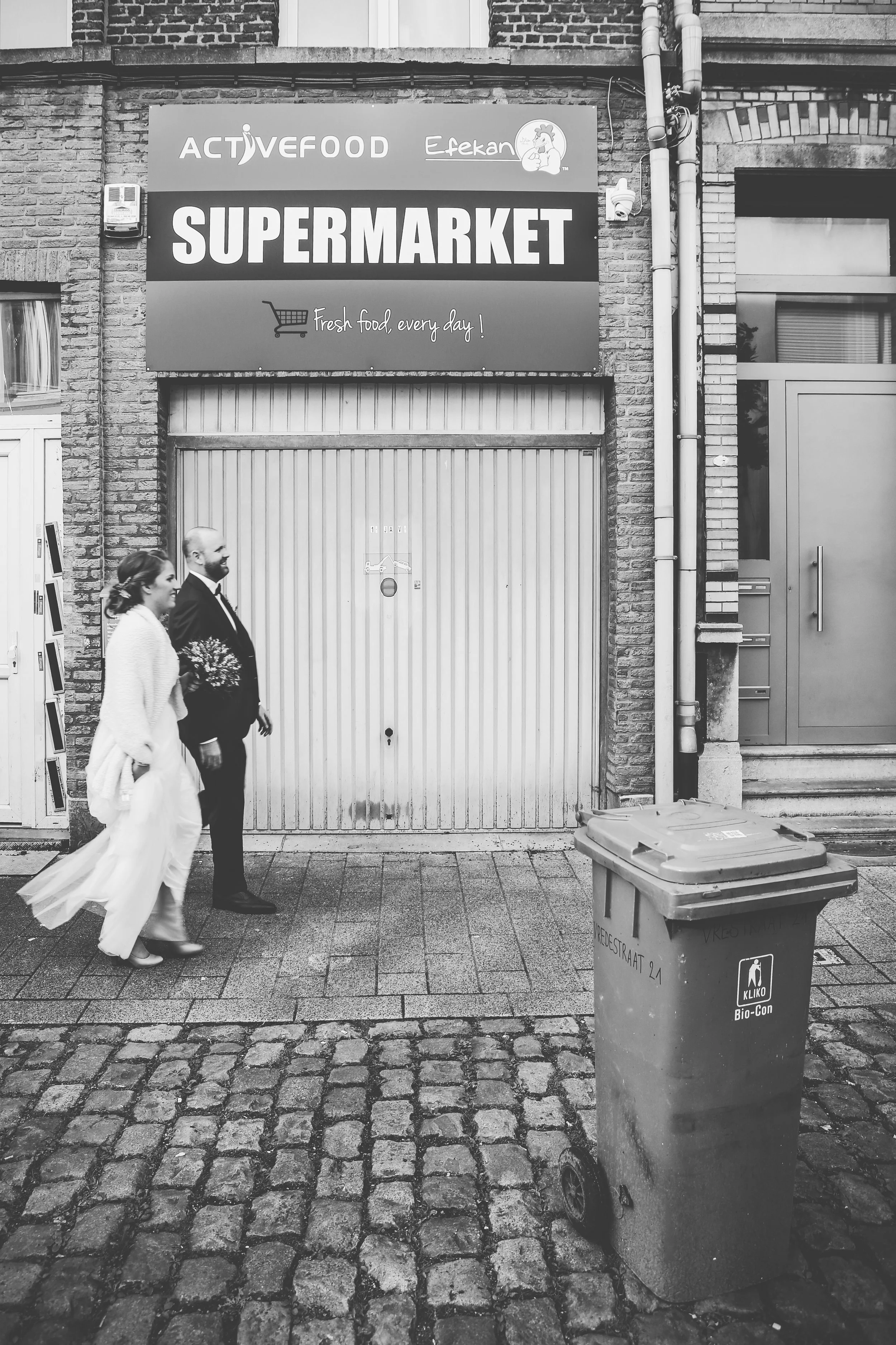 A man and woman, dressed in formal attire, walking on a cobblestone street in front of a closed storefront with a sign reading 'Supermarket' and a banner for 'Activefood Efeqan.' The woman is holding flowers, and the man is smiling. A large trash bin
