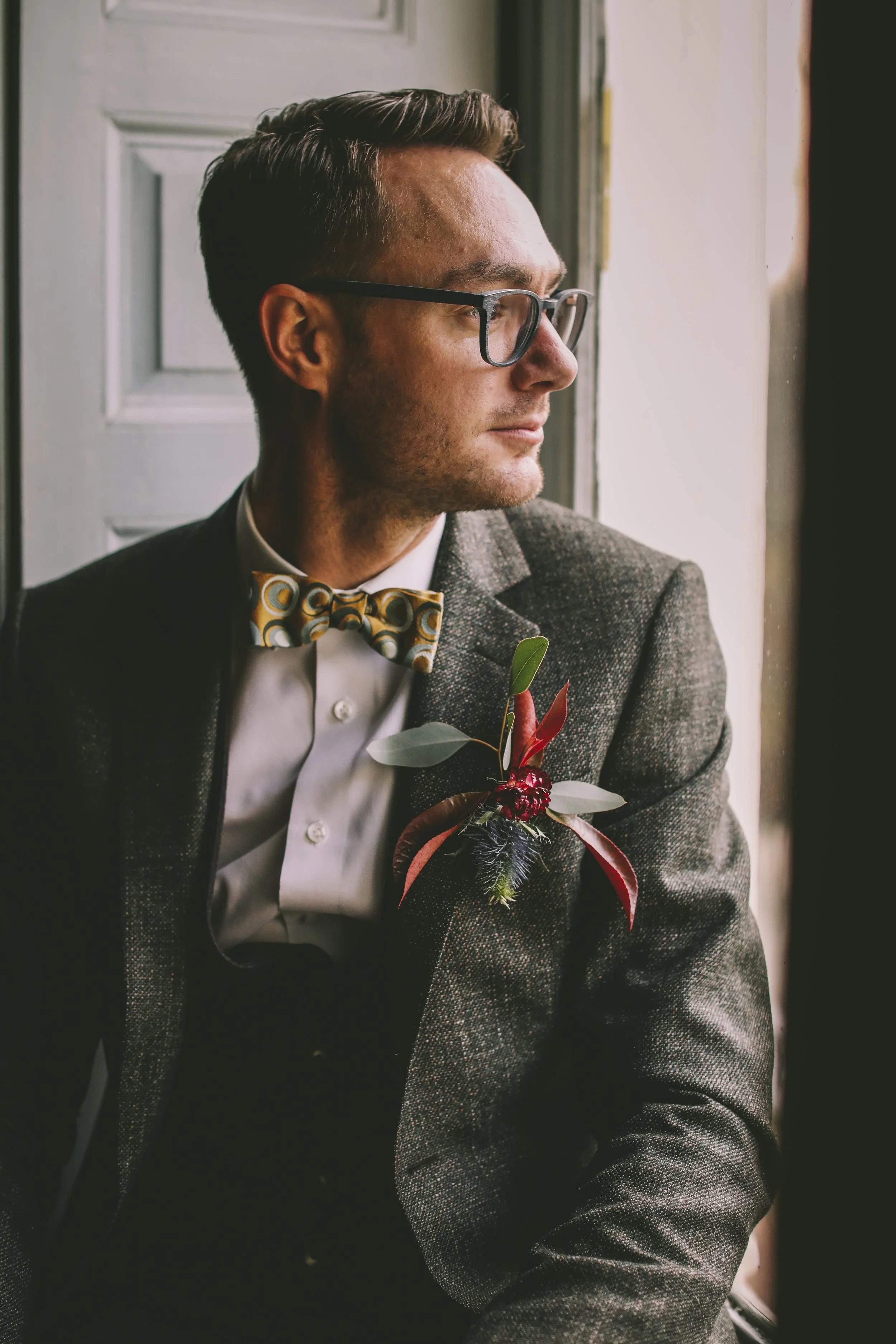 A man with glasses and styled hair, dressed in a suit with a yellow patterned bow tie, looking out of a window with a boutonniere featuring greenery and red elements on his lapel.