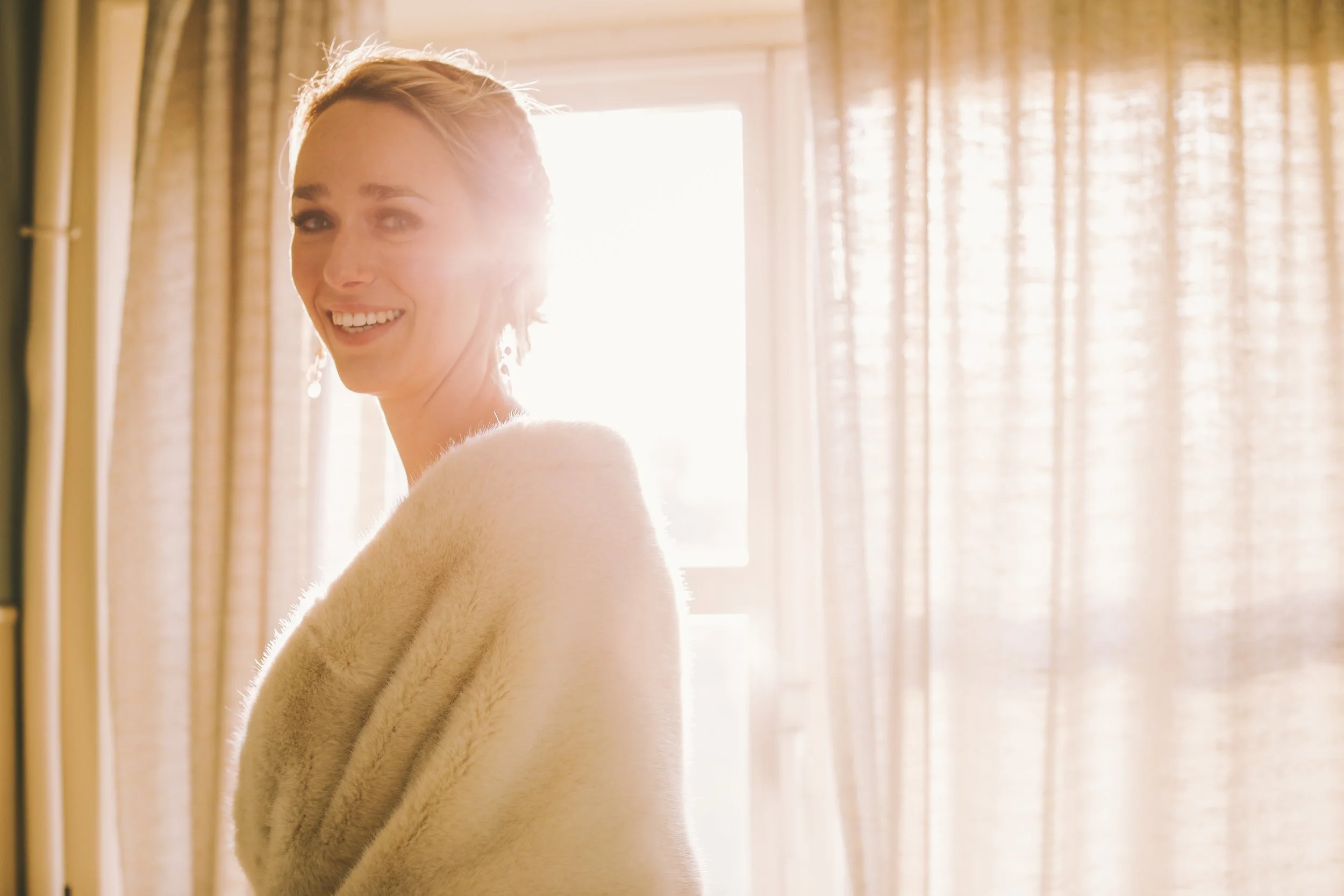 A woman with short blonde hair smiling in front of a bright window, wearing a cream-colored fur coat.