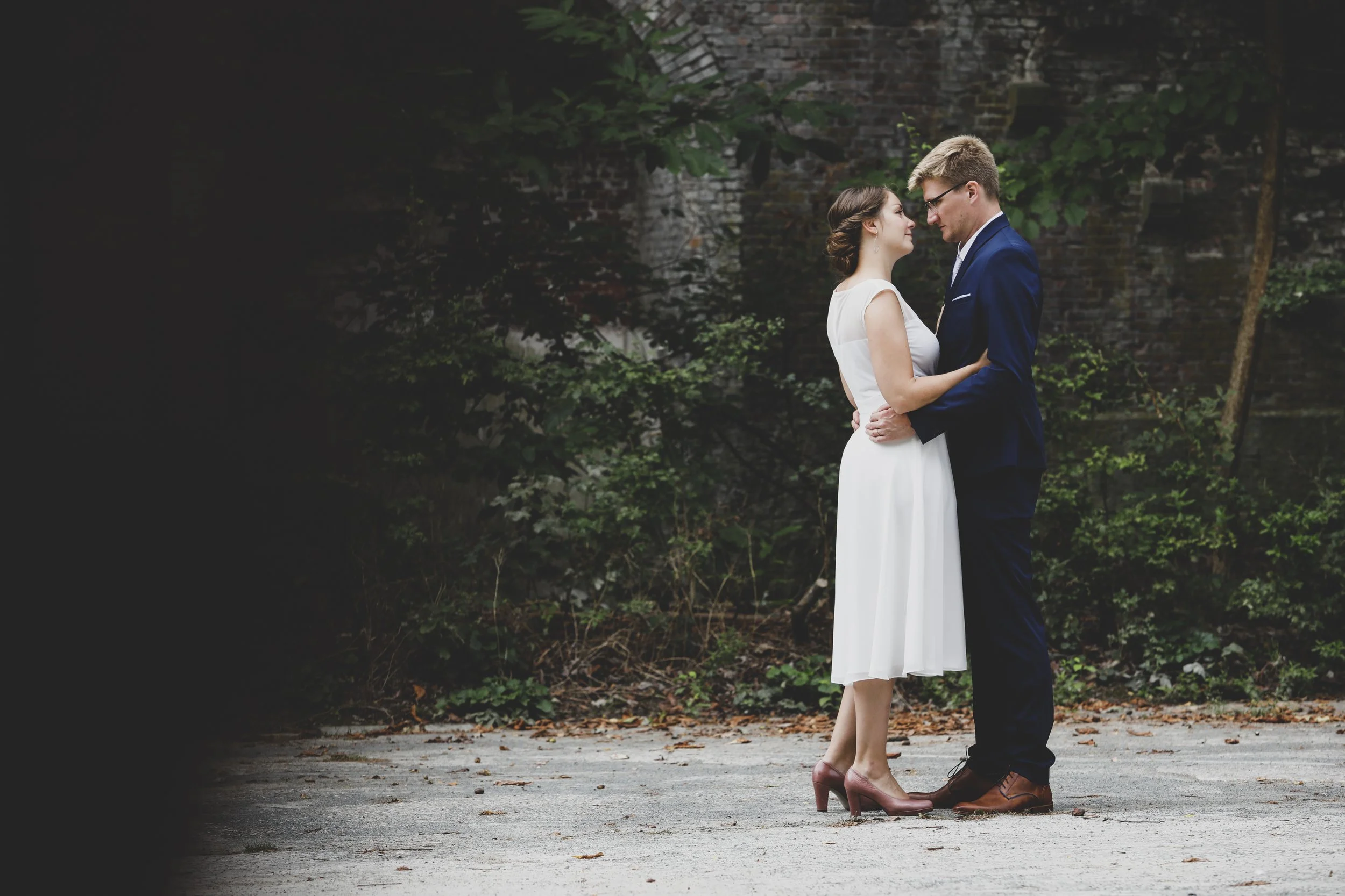 A young couple dressed in wedding attire embracing outdoors against a backdrop of green bushes and a brick wall, standing on a gravel surface.