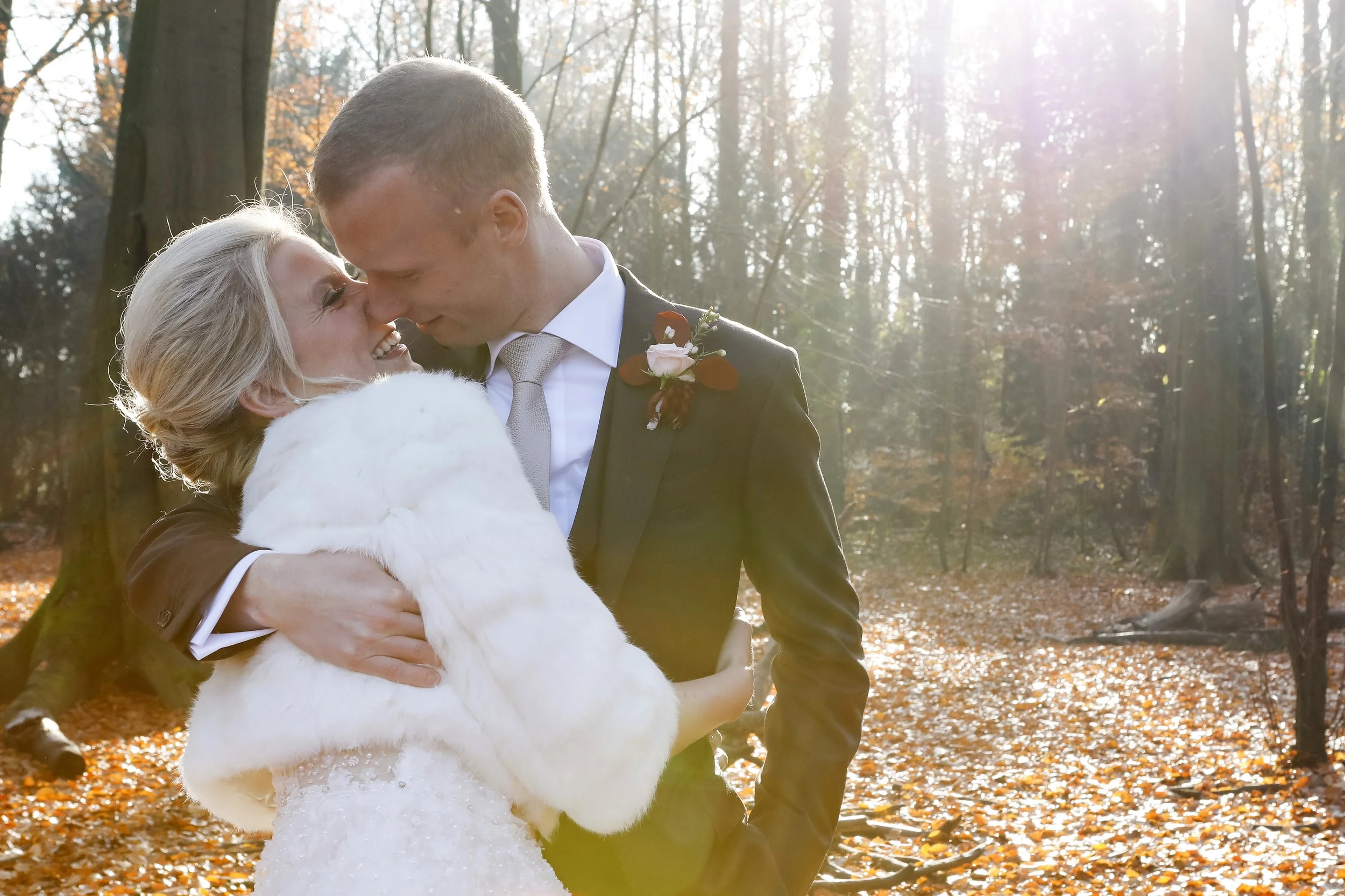 A bride and groom embracing and smiling in a wooded area during autumn, with sunlight filtering through the trees.
