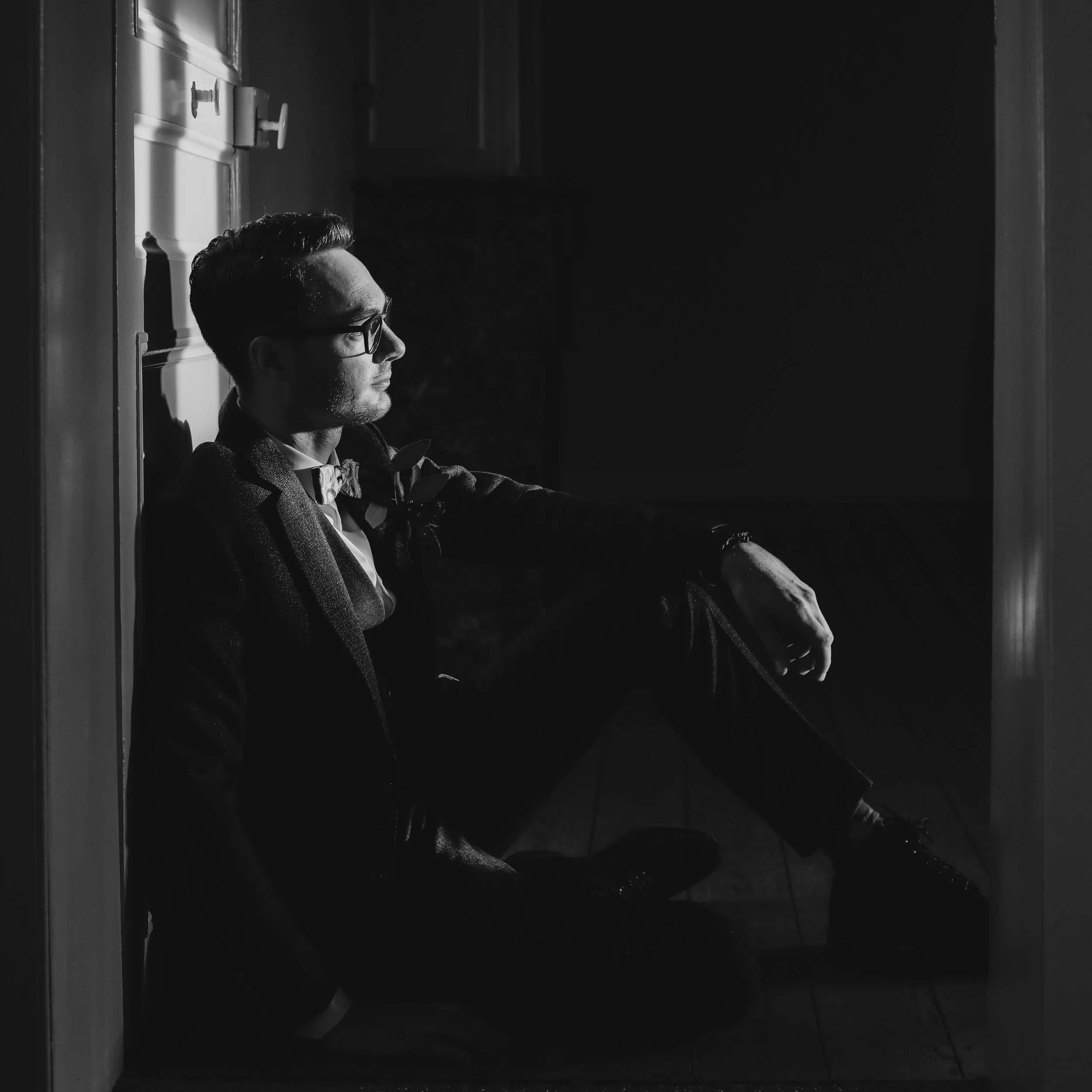 A man sitting on the floor in a dark room with a side profile view. He is wearing glasses, a blazer, and a bowtie, with light highlighting his face and hand. The background is mostly dark.