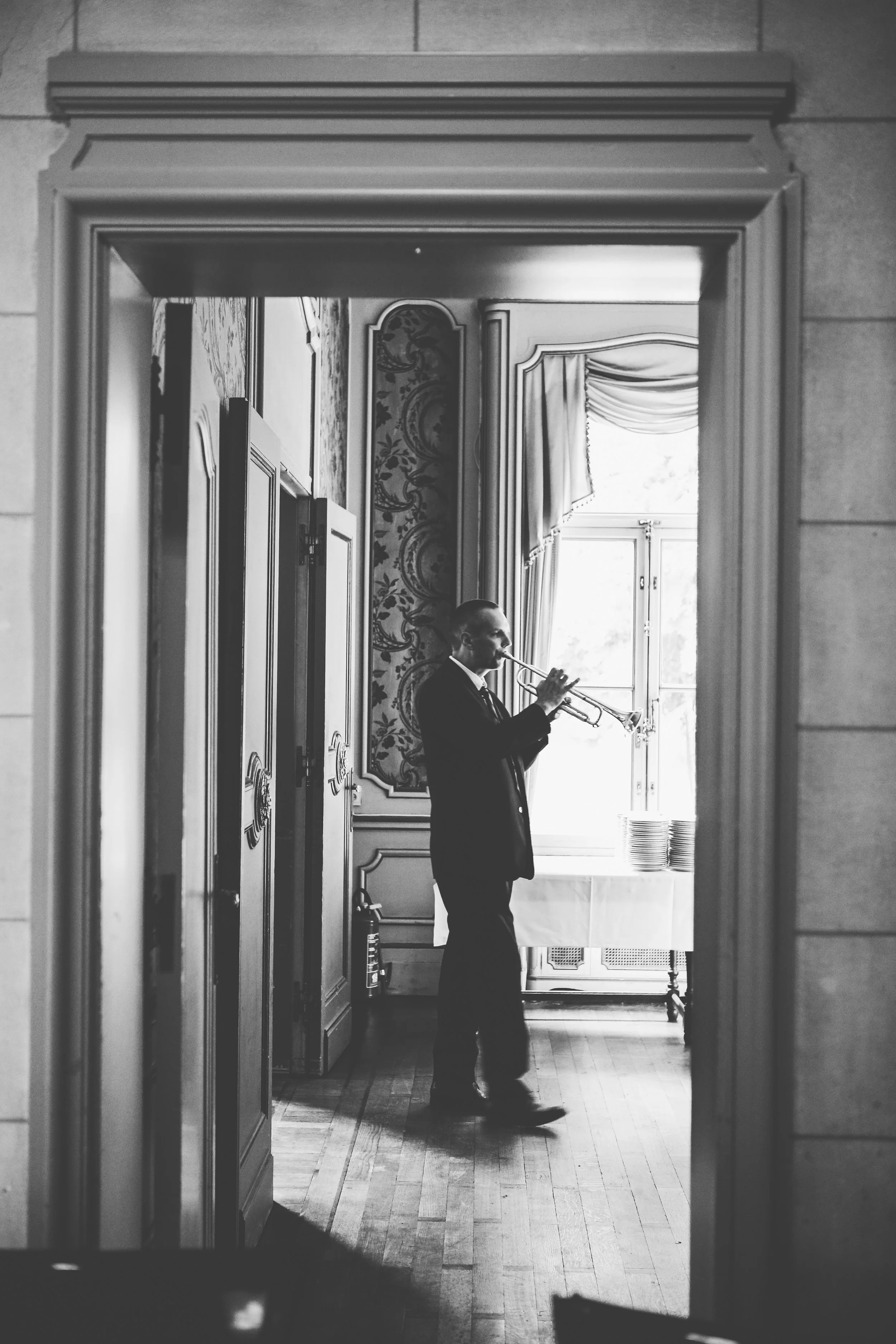A man in a suit playing trumpet indoors, viewed through a doorway with ornate trim, with sunlight coming through a window in the background.