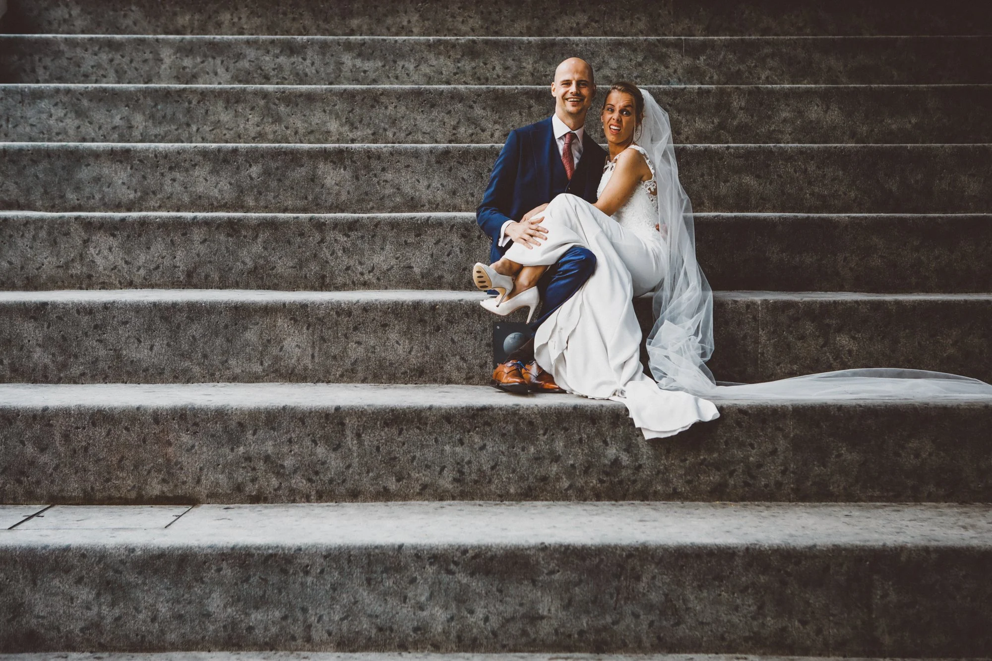 A bride and groom sitting on stone stairs, the bride is wearing a white wedding dress with a veil and the groom a navy suit with a maroon tie. They are smiling and holding each other.