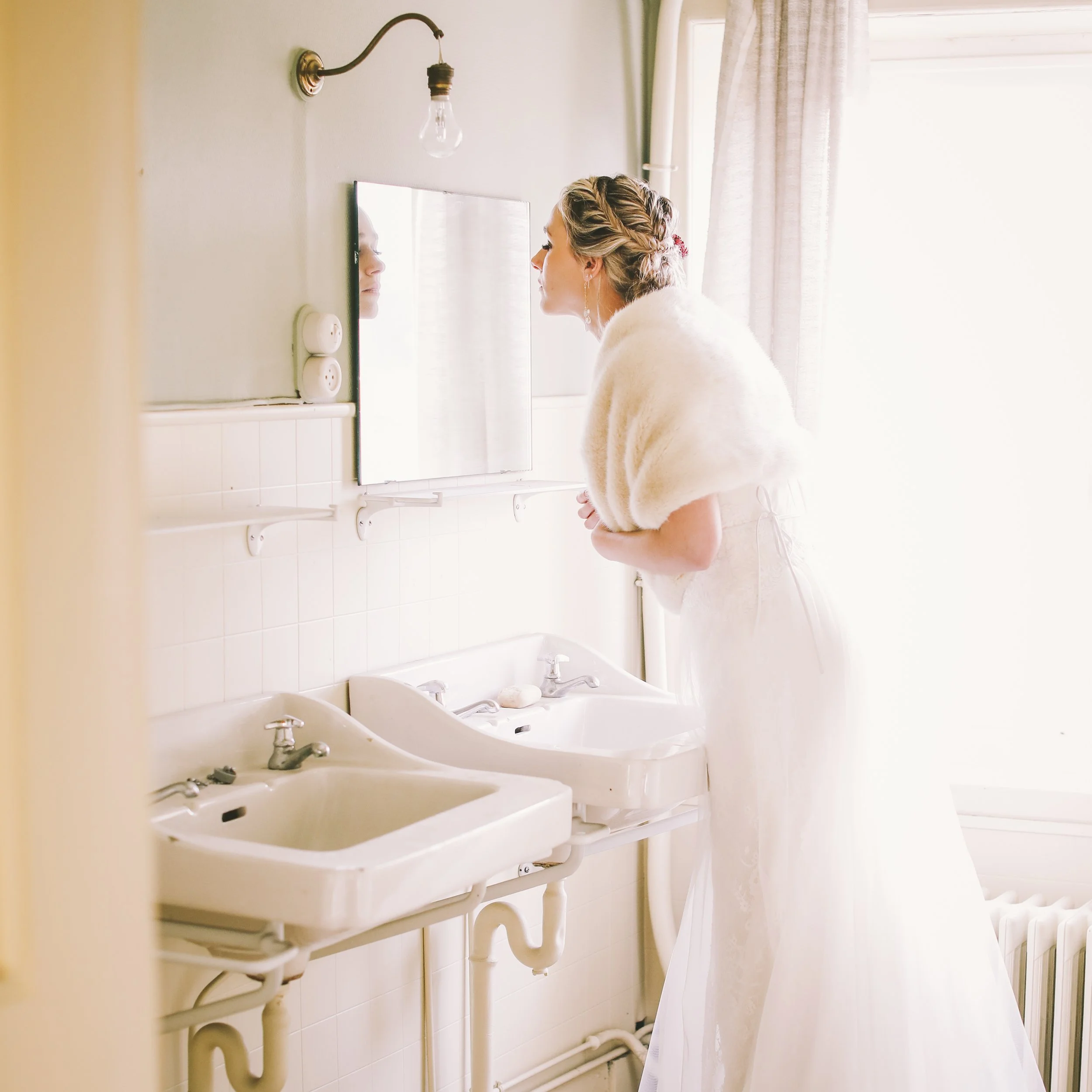 A woman in a white bridal gown and shawl looks into a mirror in a bright, softly lit bathroom.