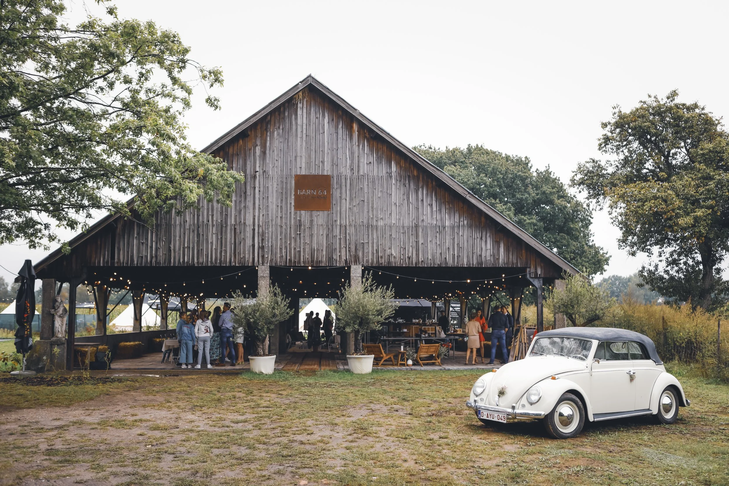 A rustic wooden barn with string lights and people socializing outside, with a vintage white Volkswagen Beetle parked on the grass nearby.