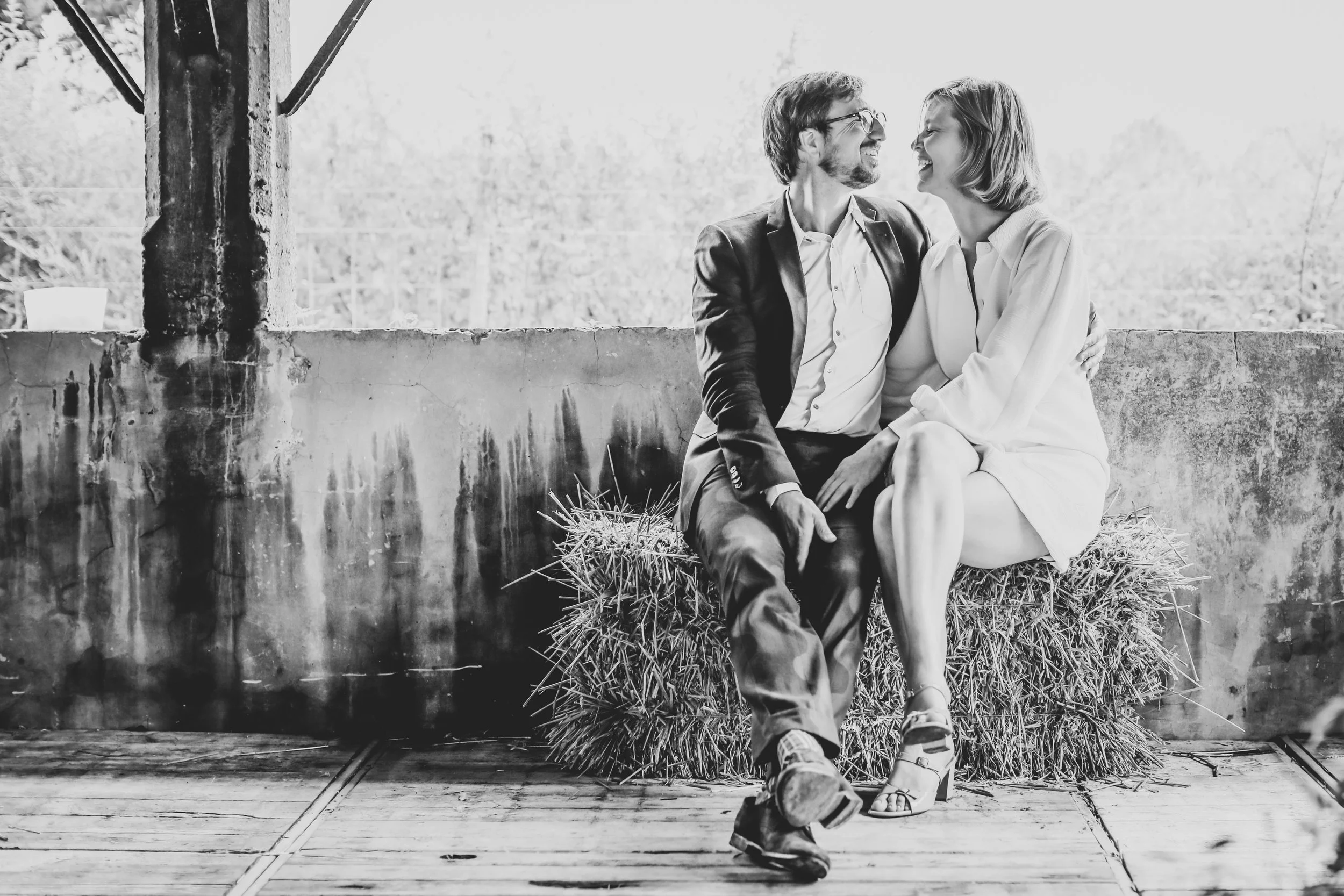 A black and white photo of a smiling couple sitting close together on a hay bale, looking at each other affectionately.