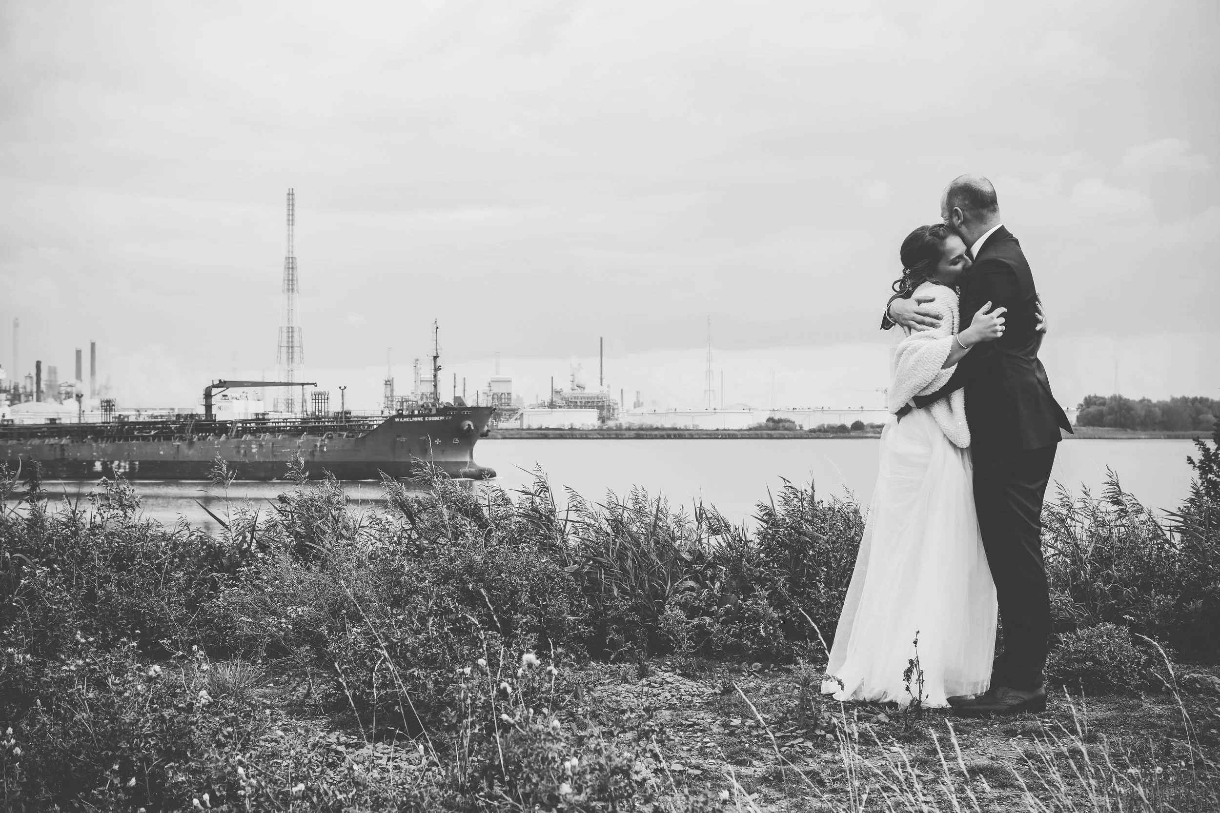 A black and white photo of a couple hugging near a river with an industrial ship and factory in the background.