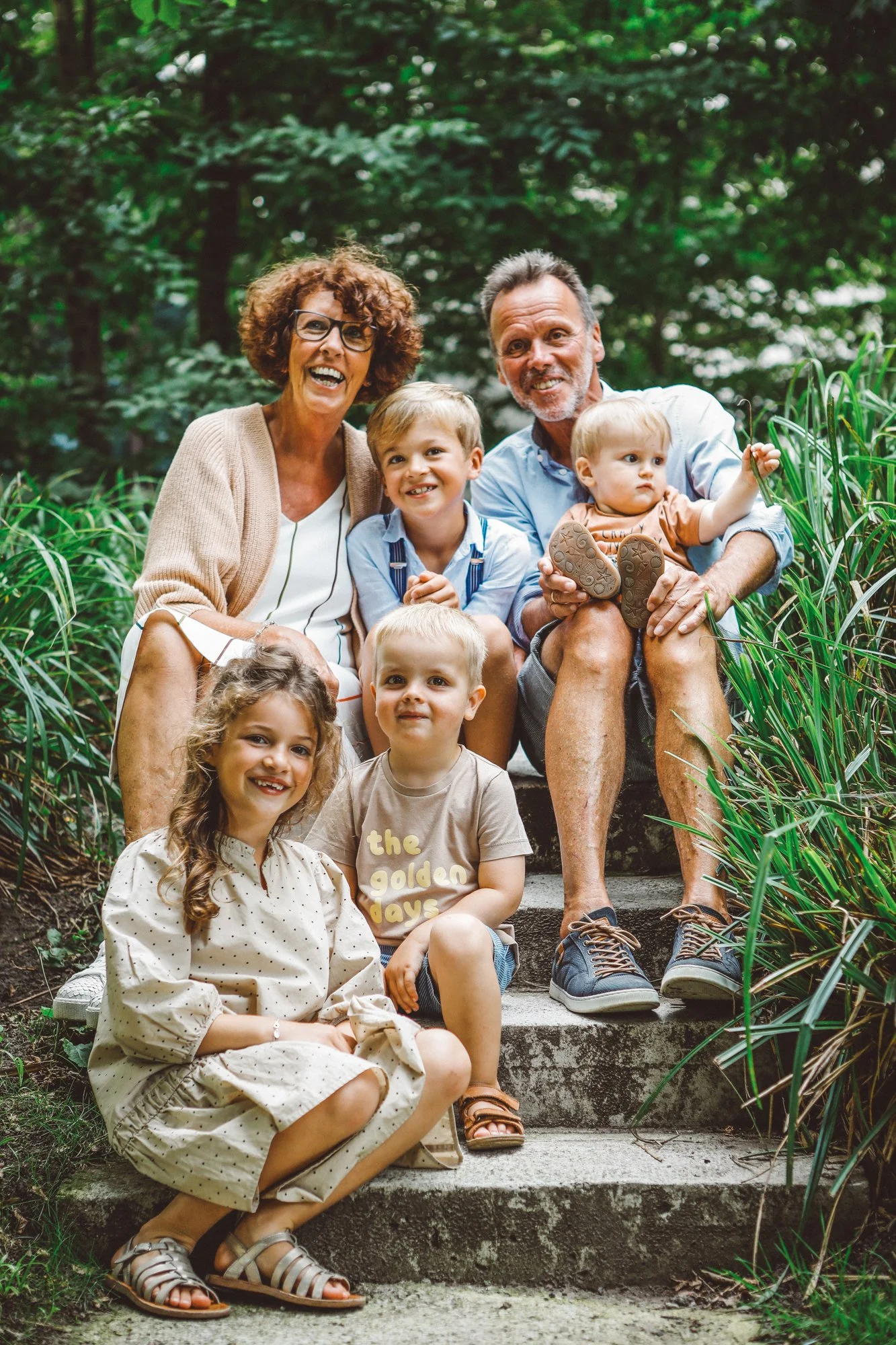 Happy multigenerational family sitting on outdoor stairs surrounded by greenery, smiling at the camera.