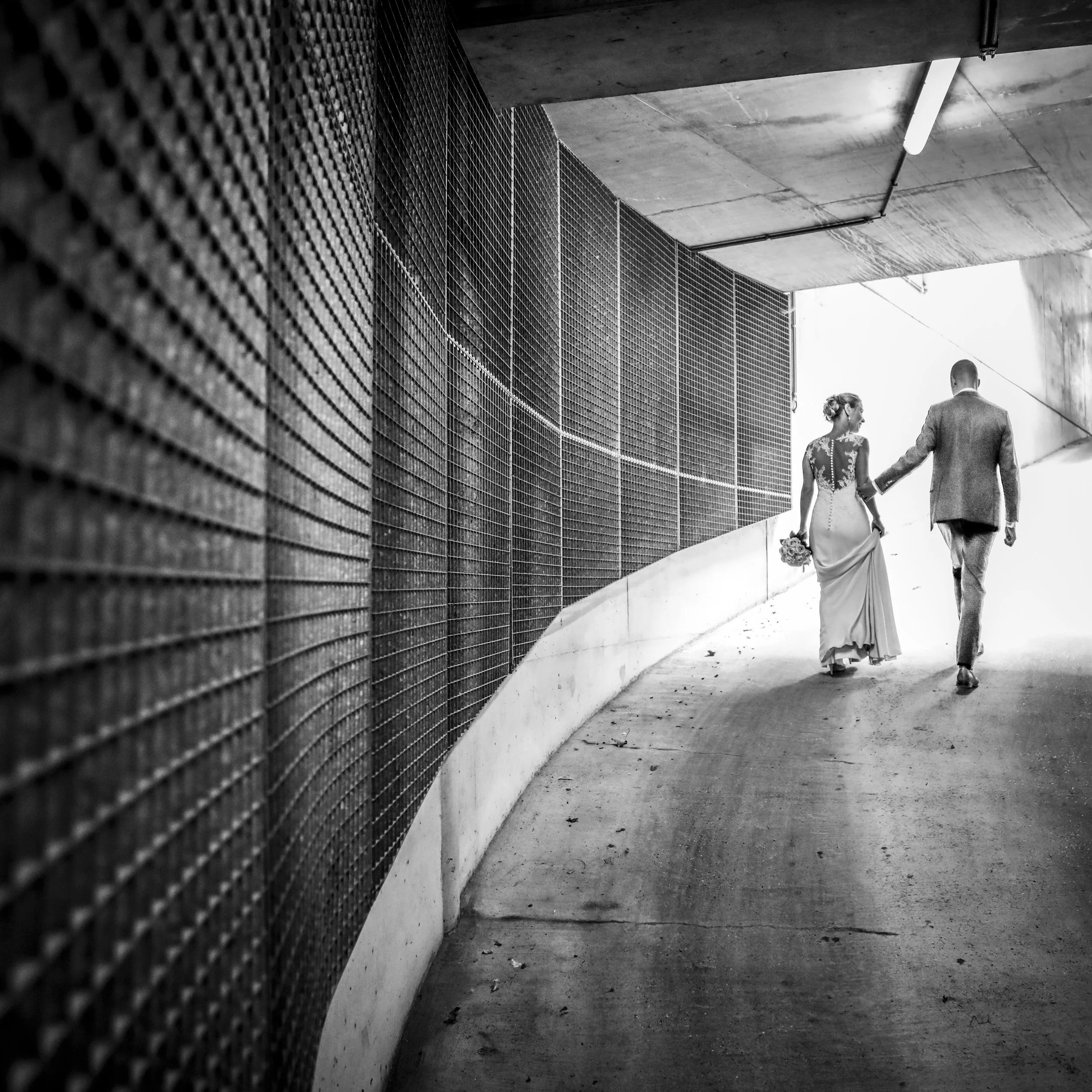 A black and white photo of a couple holding hands, walking away through an urban underpass, with the woman in a long dress holding a bouquet and the man in a suit.