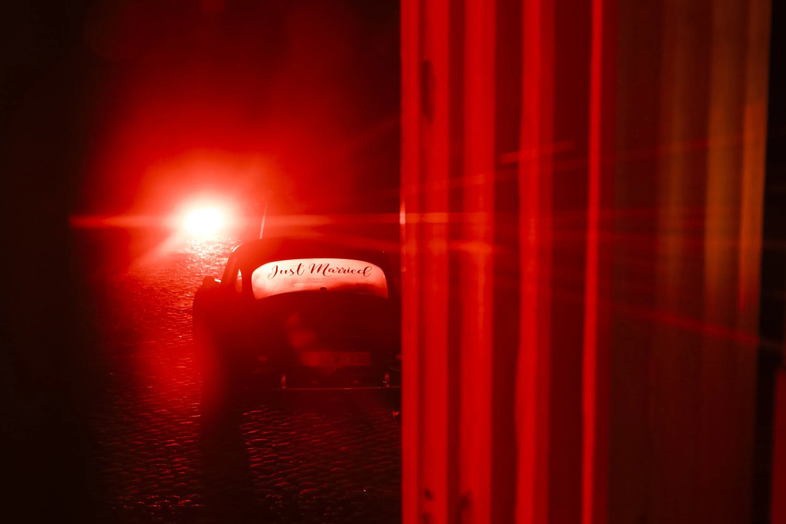 A small car with a 'Just Married' sign on the back, parked on a cobblestone street at night, illuminated by a bright red light behind it, with red bars on the right side of the image.
