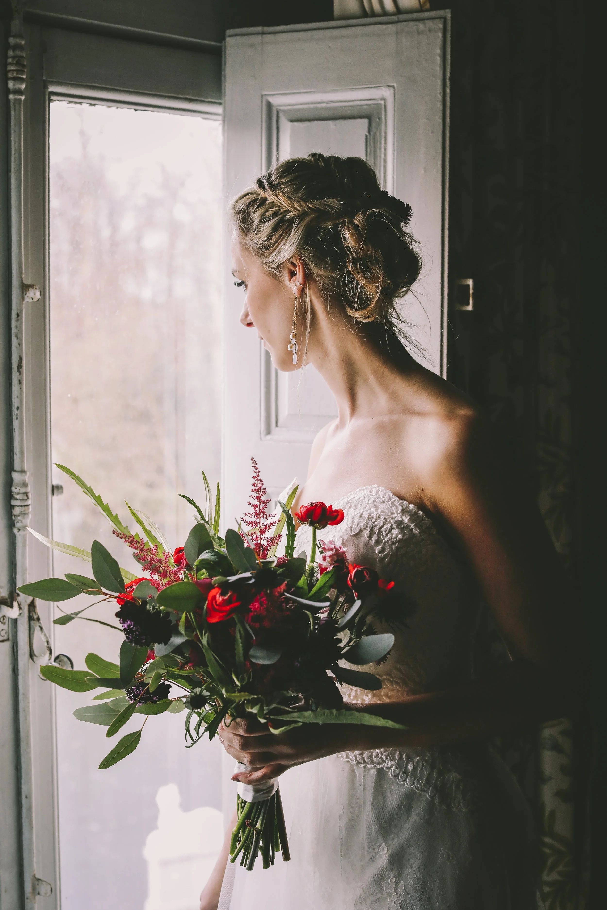 A bride with braided hair holding a bouquet of red and pink flowers, standing beside a window.