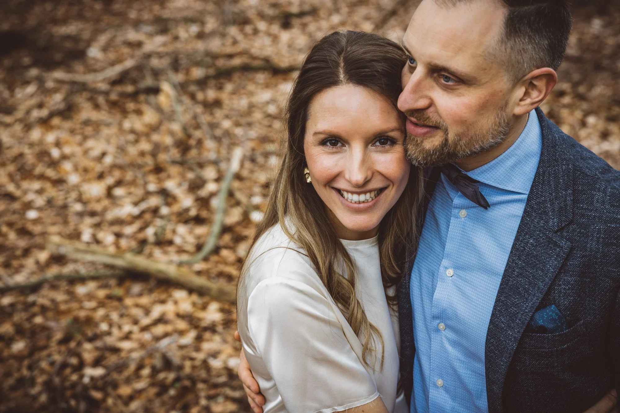 A smiling woman with long brown hair and a man with short hair and a beard standing close together outdoors on a bed of fallen leaves.