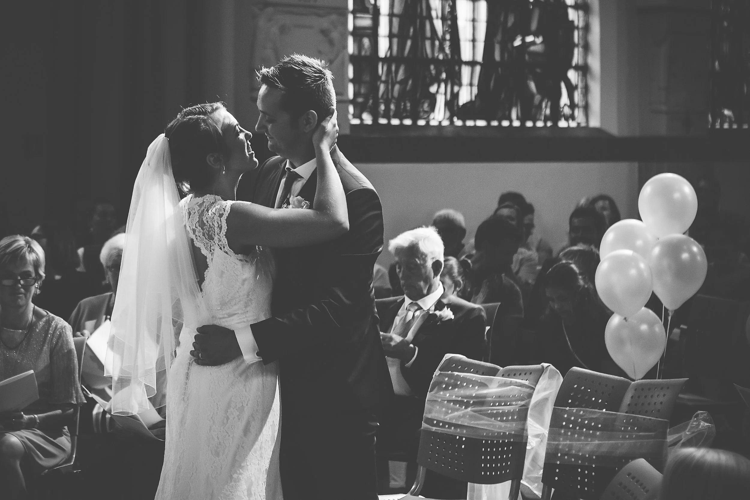 Black and white photo of a wedding dance between a bride in a lace dress and a groom in a tuxedo, surrounded by seated guests.