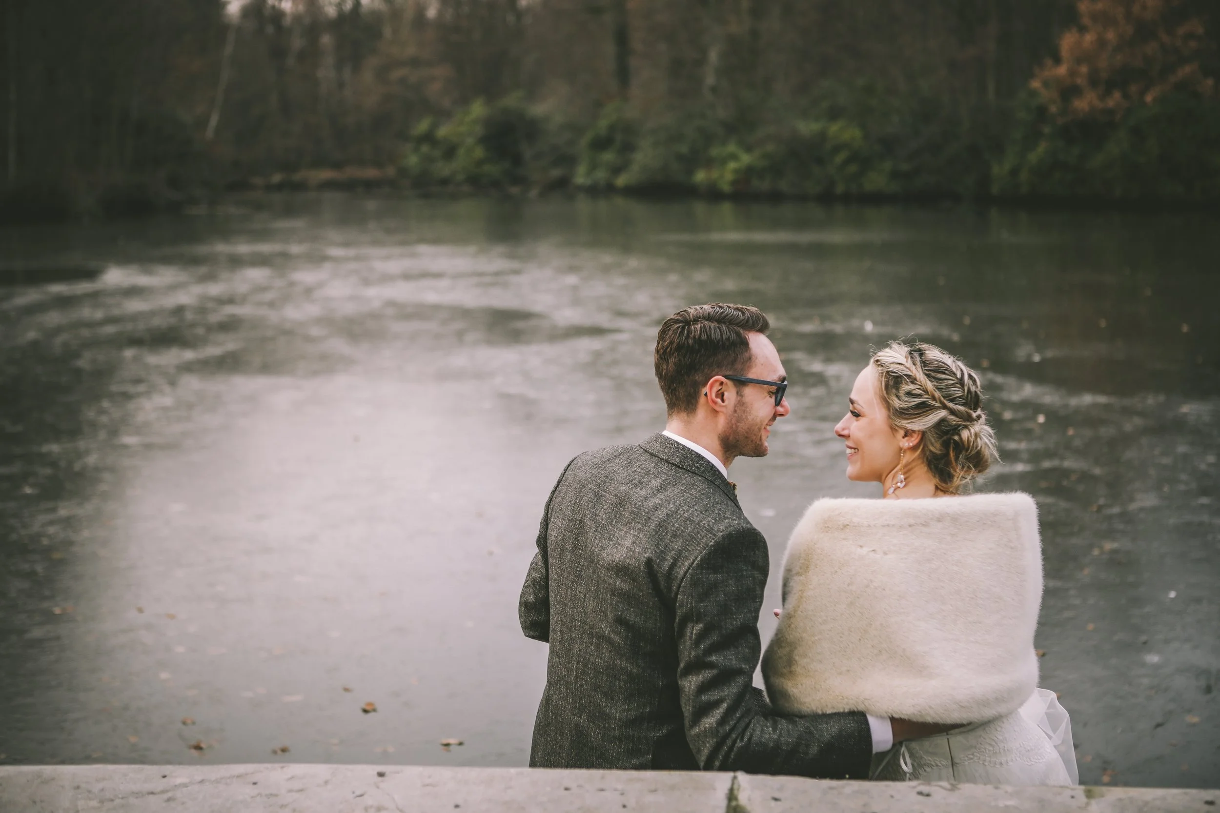 A couple is sitting by a river, smiling at each other, with trees in the background.