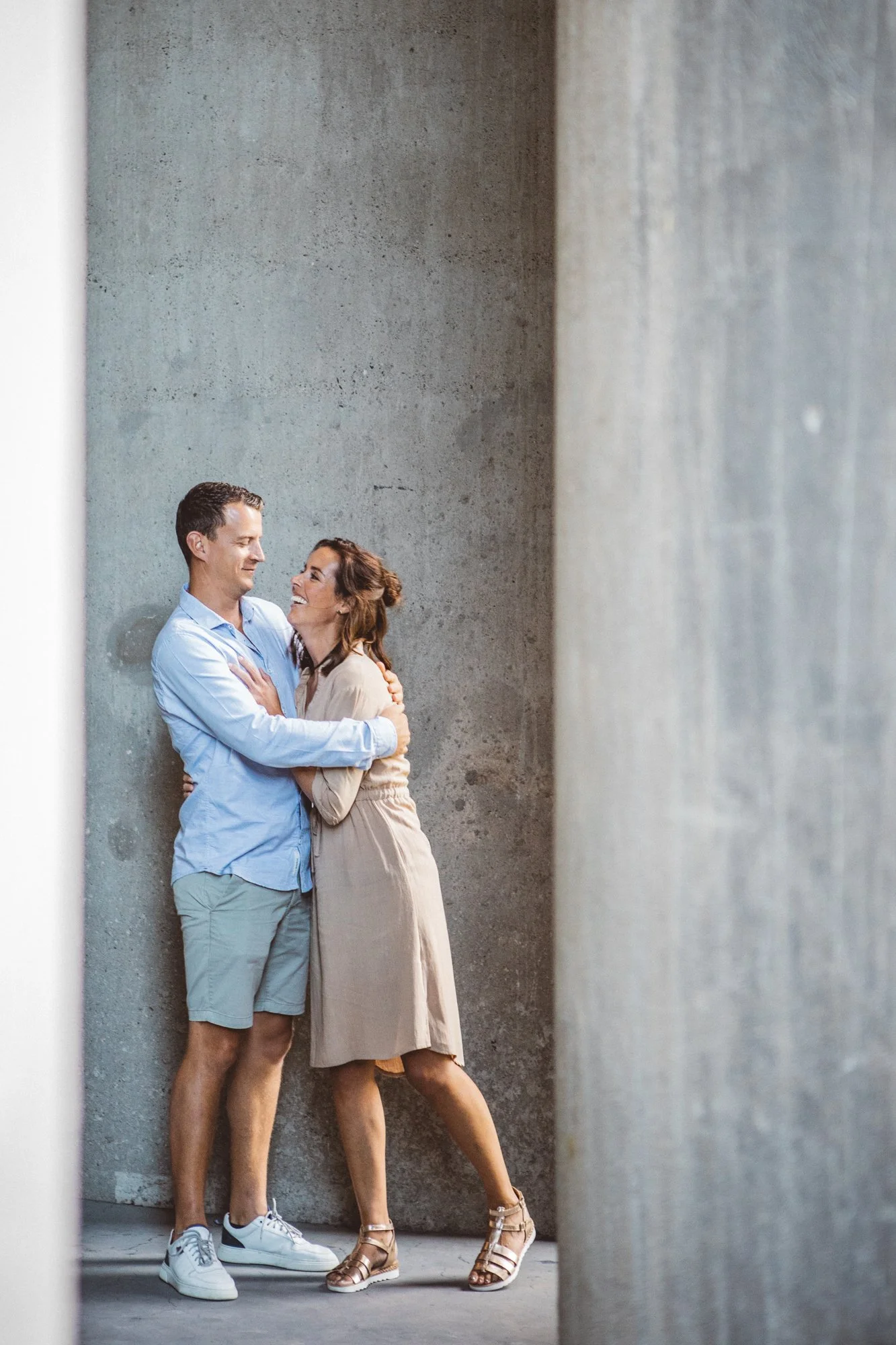 A man and woman stand close together, smiling and looking at each other, against a concrete wall, framed by two large pillars.