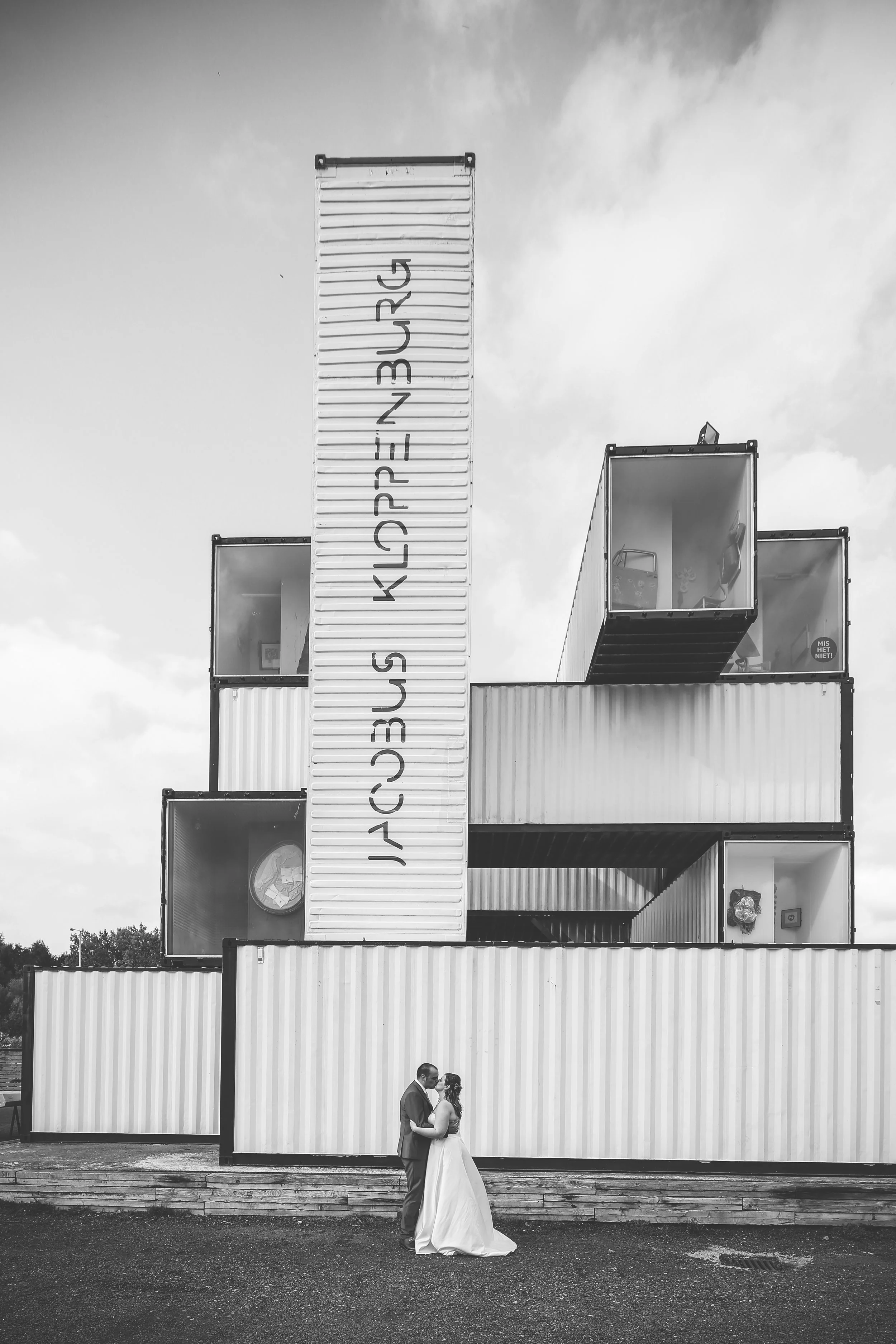 A black-and-white photo of a couple dressed in wedding attire, standing close and embracing in front of a modern building made of stacked shipping containers, with a large sign written in a language that appears to be upside down.