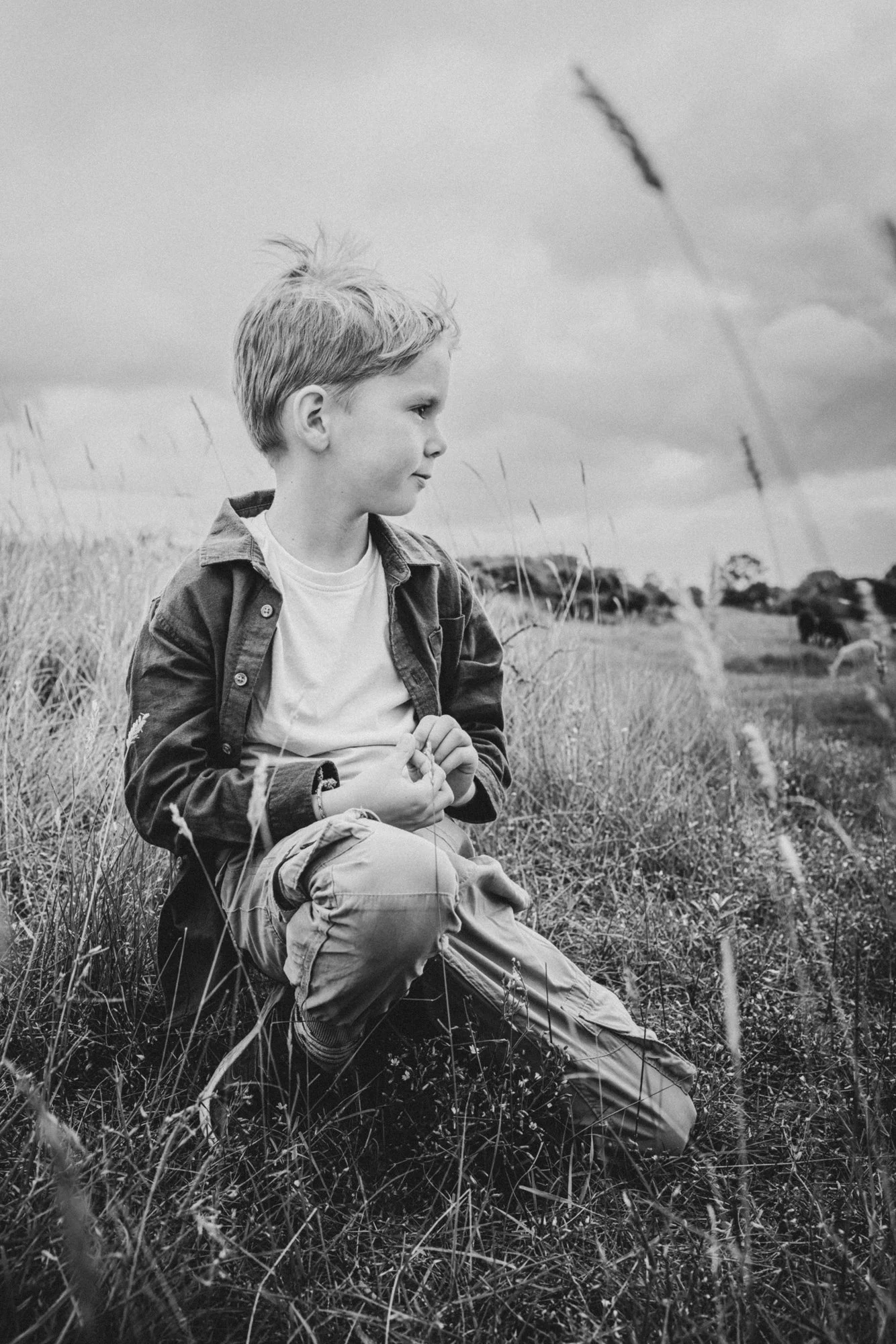 A young boy sitting in a grassy field, looking to the side, with a cloudy sky and trees in the background.