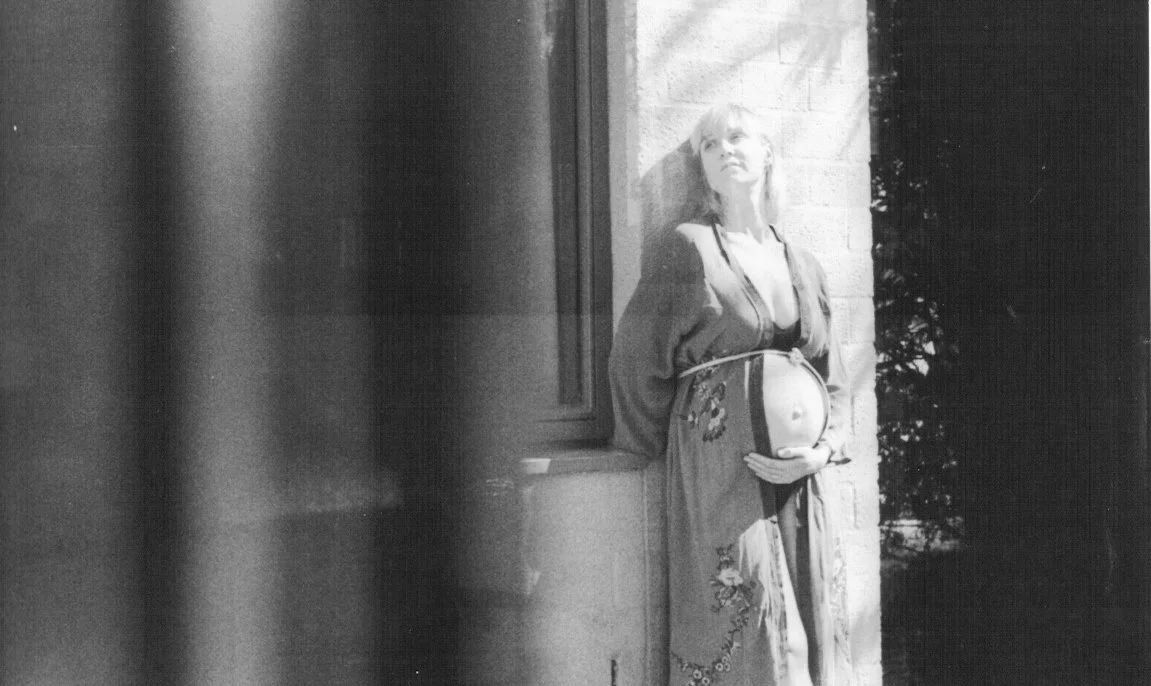 Black and white photo of a pregnant woman standing outside, leaning against a wall, with a window nearby and looking up.