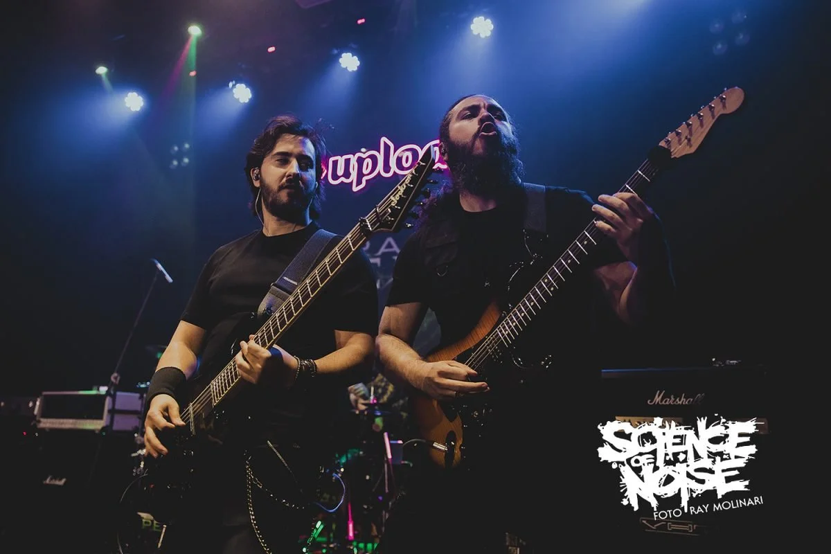 Dos músicos de la banda Rabia Perez tocando Metal moderno, groove metal, metalcore y deathcore en guitarras eléctricas en un escenario con luces de colores y un cartel que dice 'Suplon'.