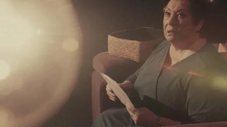 Mujer mayor sentada en un sillón con libros y sosteniendo una hoja, en un ambiente con luz tenue.Madrid. Metal moderno, metalcore, Groove metal y deathcore.