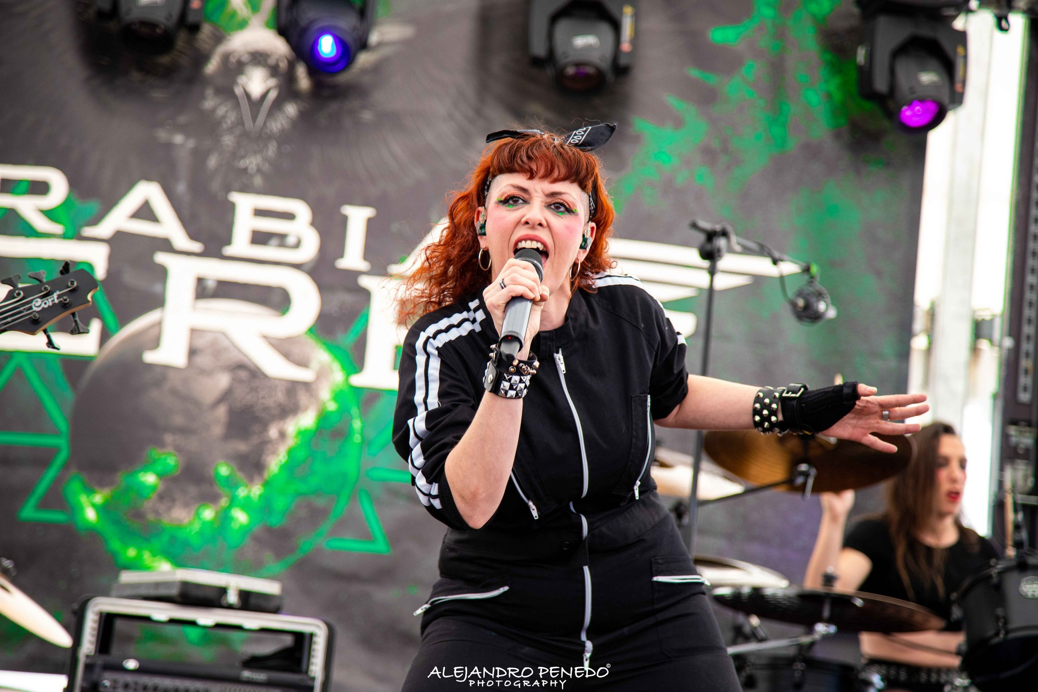 Mujer cantante con cabello rizado rojo, usando chaqueta negra con rayas blancas en las mangas, cantando Metal moderno, groove metal, metalcore y deathcore en un escenario de madrid durante concierto, con fondo decorativo y banda musical.