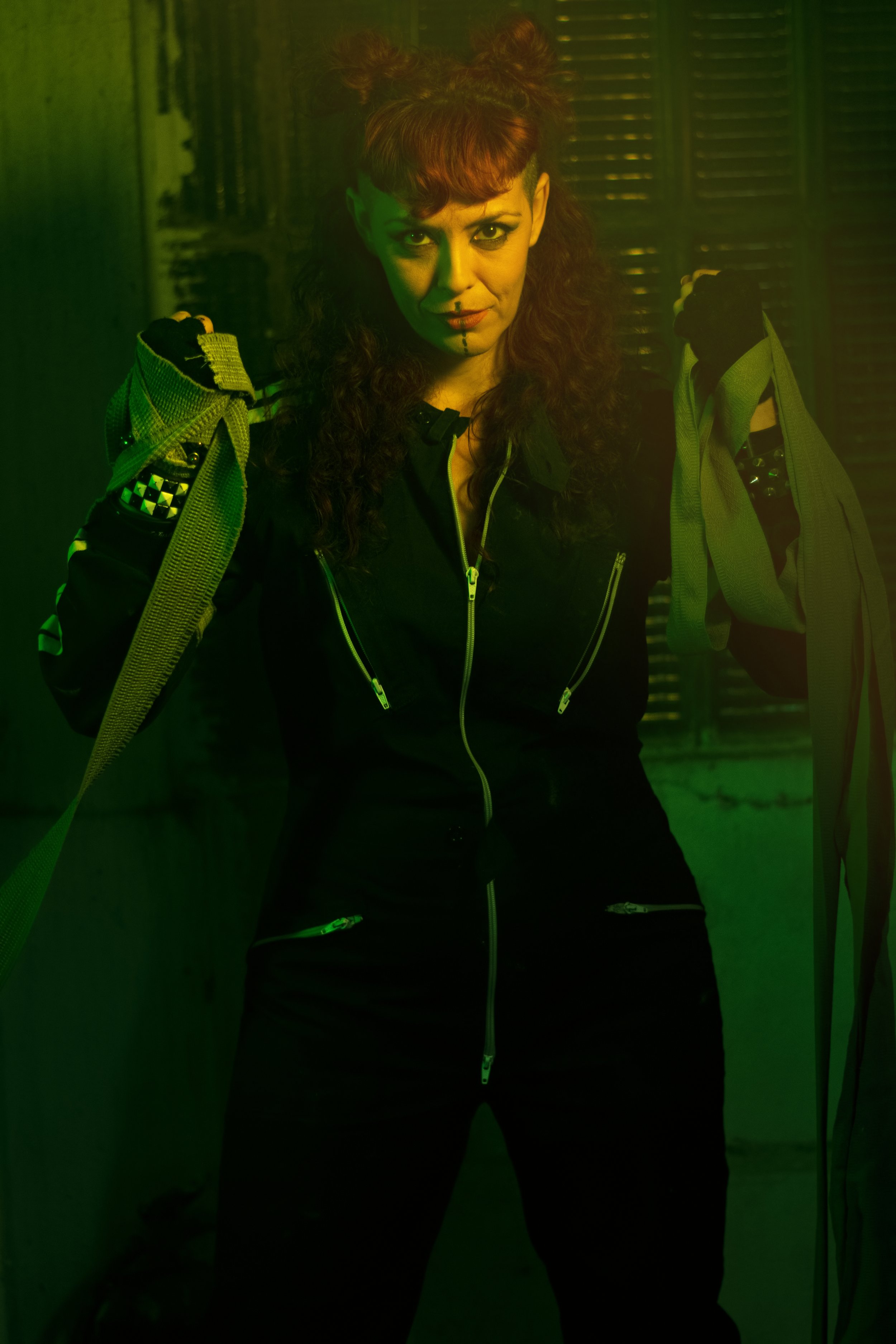 Mujer vocalista de la banda de Metal moderno, groove metal, metalcore y deathcore Rabia Perez con cabello rizado rojo y cabello en doble moño, vestida con ropa negra con cremalleras, con guantes negros y con maletas o bolsas en las manos, en un entor