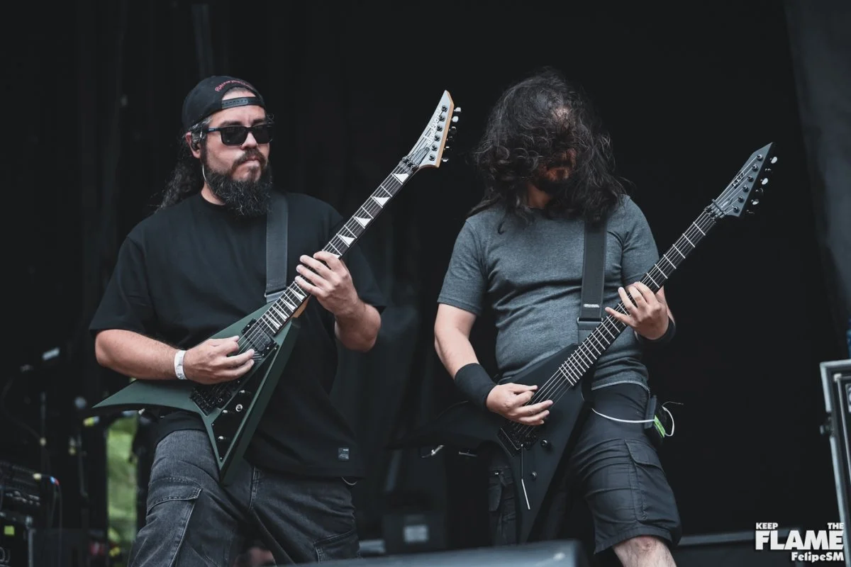Dos músicos tocando Metal moderno, groove metal, metalcore y deathcore en guitarras eléctricas en un escenario, uno con gafas oscuras y sombrero, el otro con cabello rizado y sin gafas, fondo oscuro.