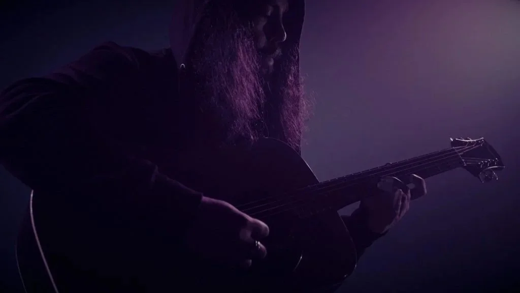 Persona tocando guitarra en un entorno oscuro con iluminación suave y de tono violeta.