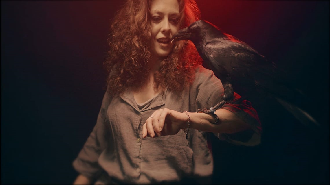 Mujer con cabello rizado, vestida con blusa gris, interactuando con un cuervo en su brazo para el rodaje de un videoclip de Metal moderno, groove metal, metalcore y deathcore en Madrid.