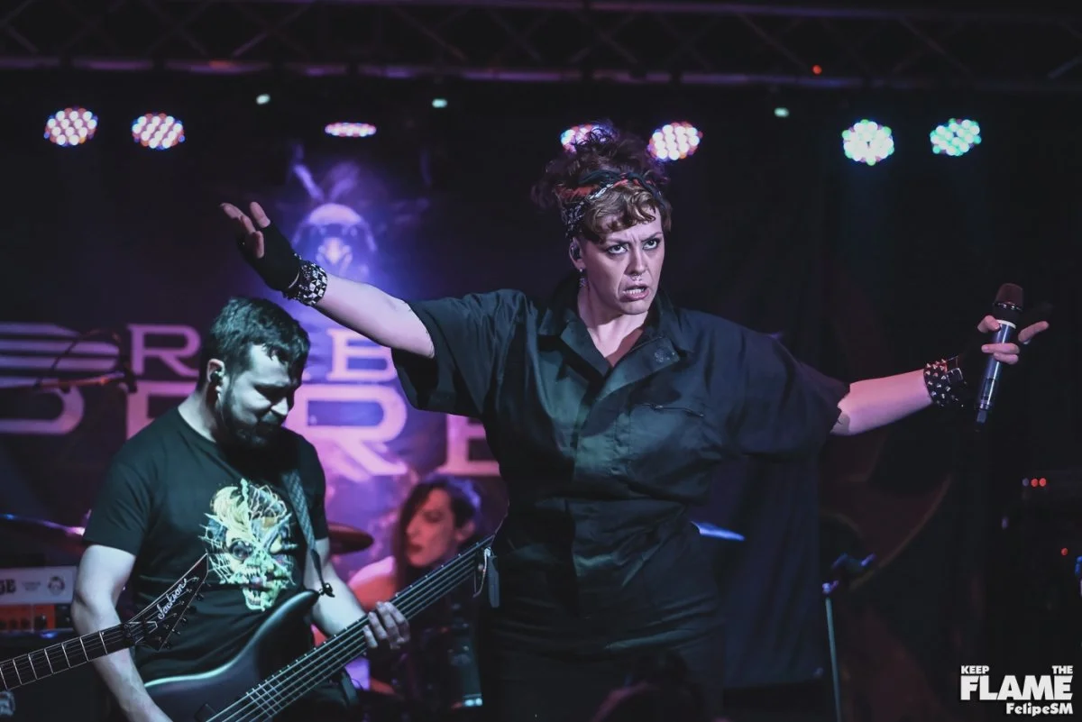 Una mujer con cabello rizado y peinado con pañuelo en la cabeza, vestida con ropa negra, en el escenario durante una presentación musical, sosteniendo un micrófono y con expresión enérgica. En el fondo, un hombre tocando la guitarra eléctrica y una mujer tocando la batería, ambos en un concierto con iluminación de escenario