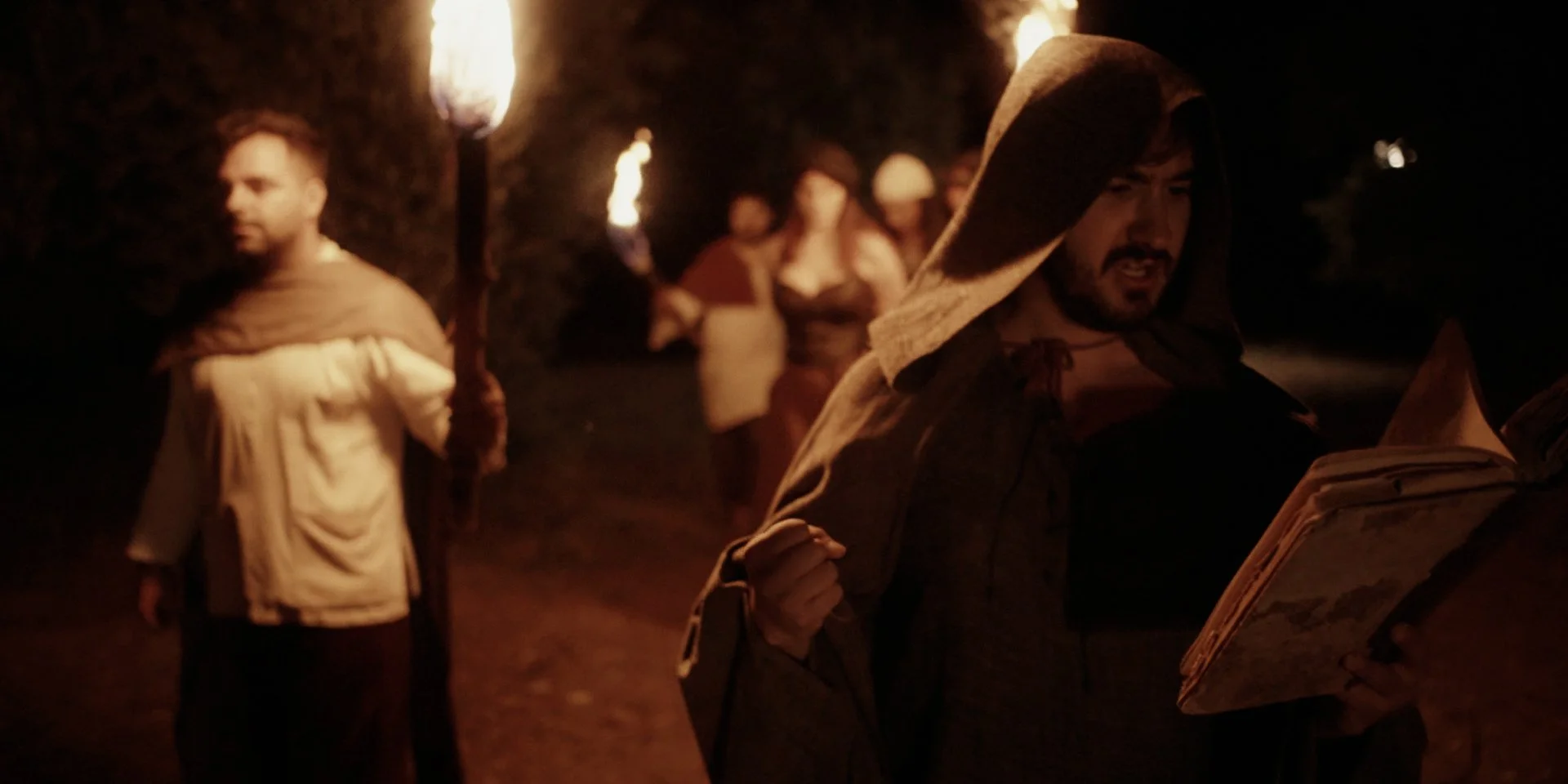 Grupo de personas en una noche con antorchas, una persona en primer plano leyendo un libro y otra en el fondo sosteniendo una antorcha.