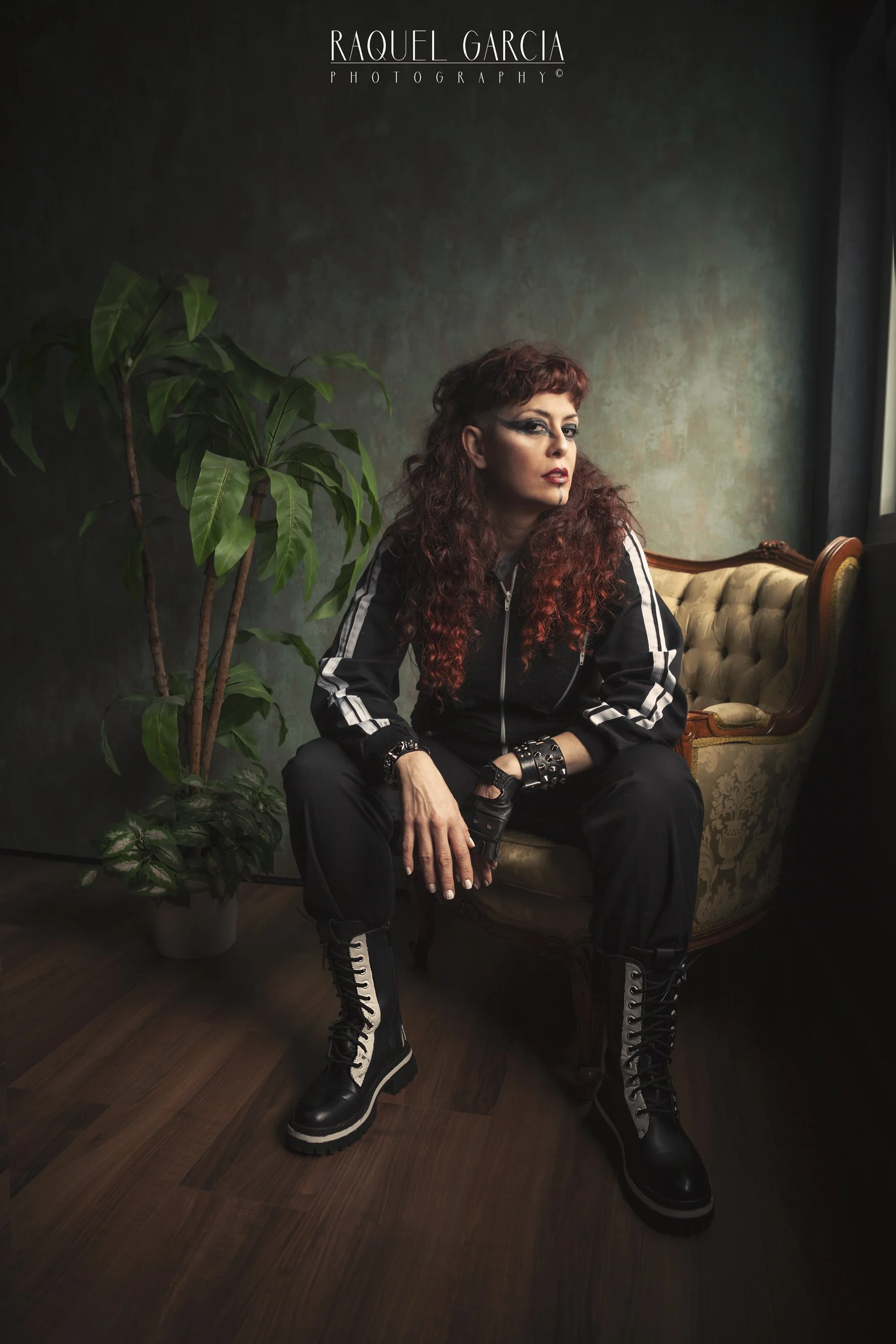Una mujer vocalista de la banda de Metal moderno, groove metal, metalcore y deathcore Rabia Perez con cabello rizado y rojo, vestida con ropa negra con detalles blancos, sentada en una silla antigua tapizada, junto a una planta decorativa, en una hab