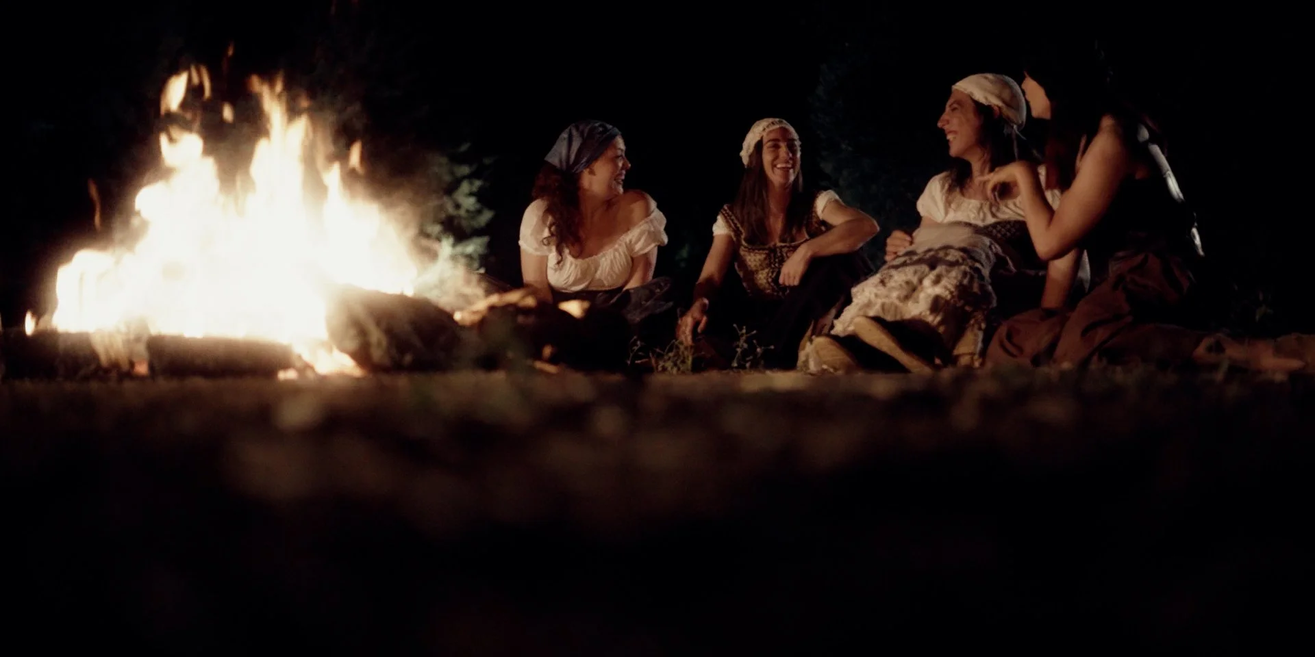 Cuatro mujeres con vestimenta antigua sentadas alrededor de una fogata en la noche, riendo y conversando.