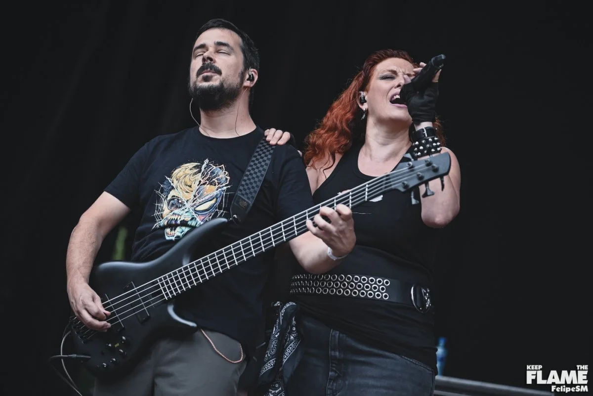 Un hombre y una mujer peliroja tocando en un concierto de Metal moderno, groove metal, metalcore y deathcore con la mujer cantando y el hombre tocando el bajo eléctrico. Ambos parecen emocionados y concentrados en su actuación.