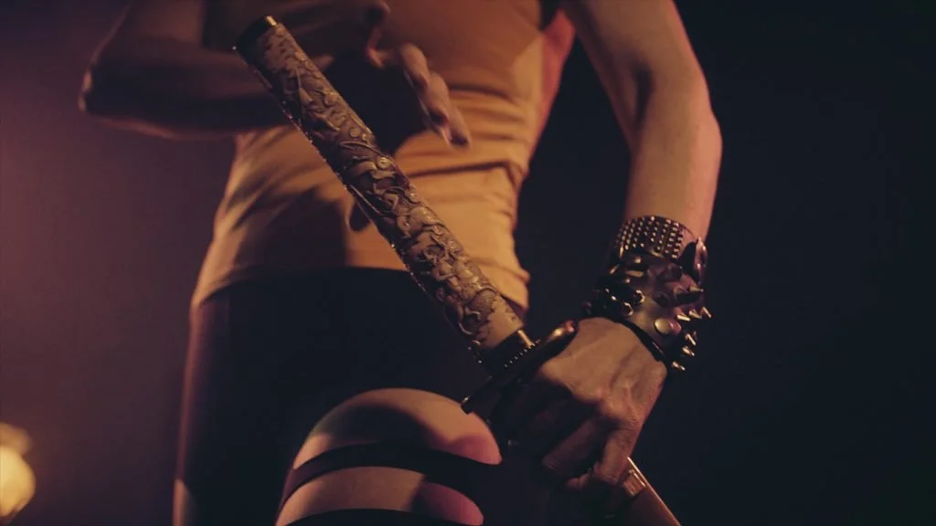 Persona sosteniendo un látigo decorado con flores, usando pulseras y un reloj de pulsera, vestida con una camiseta ajustada y pantalones cortos en un ambiente oscuro.