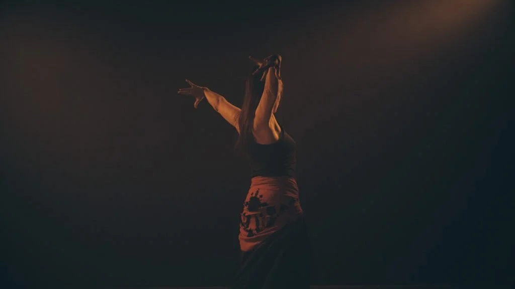 Figura humana en movimiento en un escenario oscuro, con brazos extendidos y cabello largo, vestida con ropa negra y naranja.