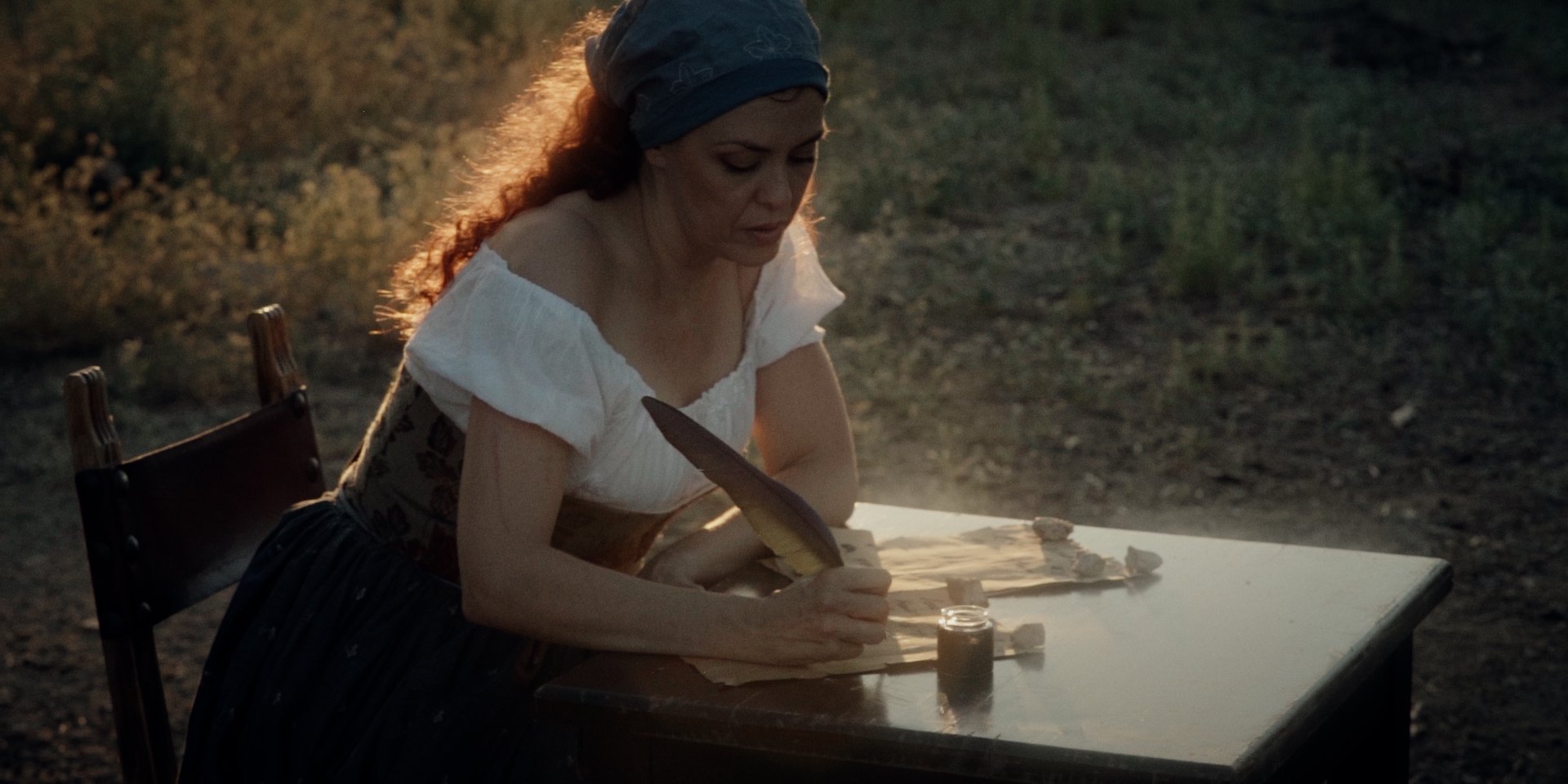 Mujer vestida con ropa de época, sentada en una silla, trabajando con plumas y tinta en una mesa en el exterior.