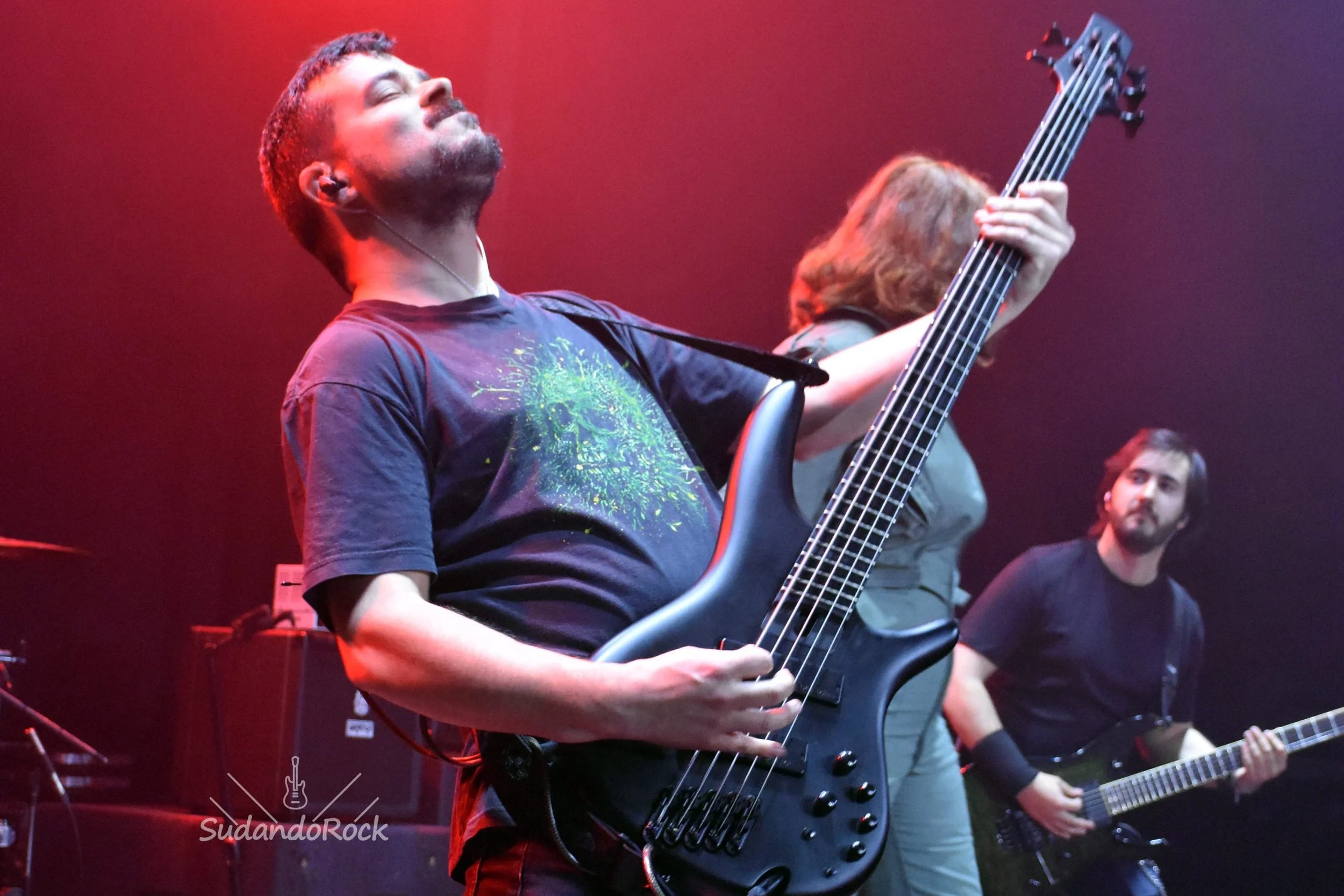 Tres músicos de la banda de Madrid Rabia Perez tocando Metal moderno, groove metal, metalcore y deathcore en un escenario, uno con guitarra eléctrica, otro con bajo y un tercero con guitarra eléctrica en el fondo, con iluminación de colores rojos y m