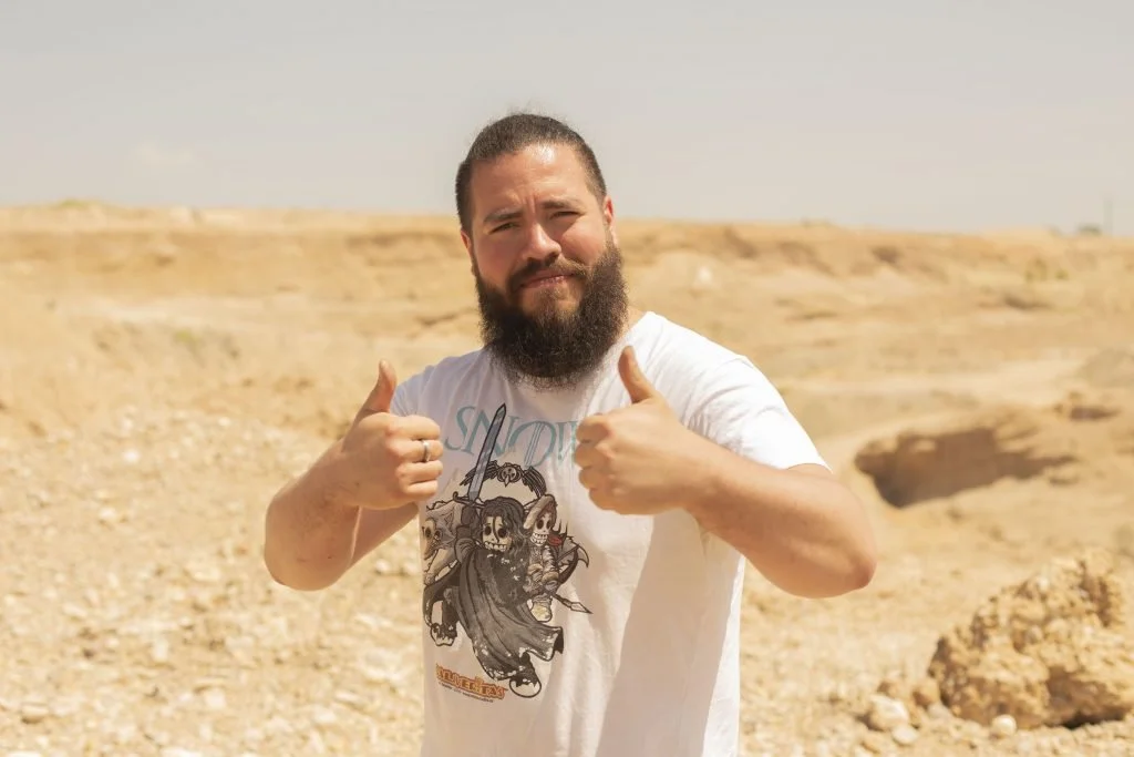 Hombre con barba y camiseta blanca en un paisaje de desierto, haciendo señal de pulgar arriba con ambas manos.