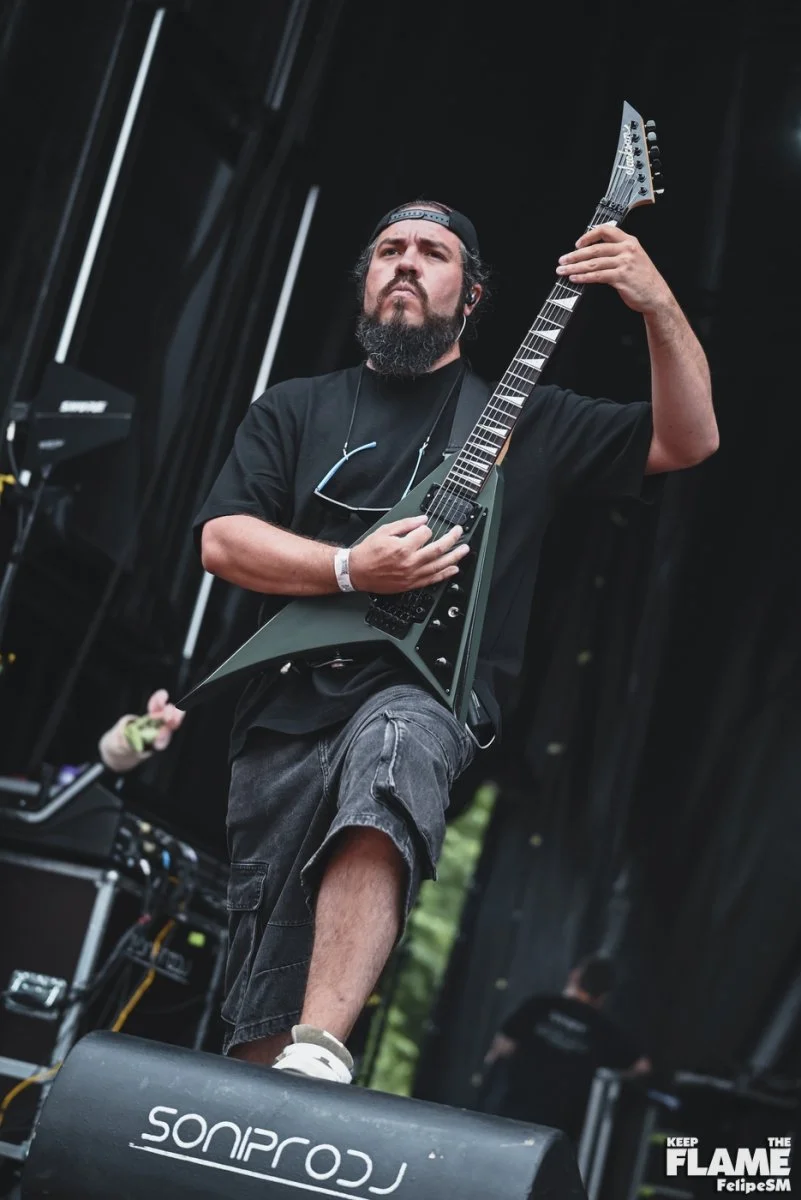 Un hombre tocando Metal moderno, groove metal, metalcore y deathcore en una guitarra eléctrica jackson en un escenario, lleva una camiseta negra, pantalones cortos oscuros, gafas colgando del cuello y usa una gorra. Está concentrado en su interpretac