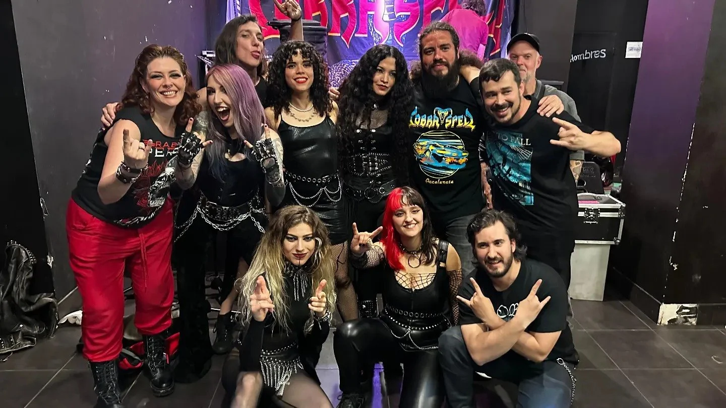 Grupo de personas en un ambiente oscuro con ropa de estilo rock y Metal moderno, groove metal, metalcore y deathcore, algunos con cadenas, maquillaje oscuro y peinados llamativos, posando y sonriendo frente a una pared con letras en colores brillante