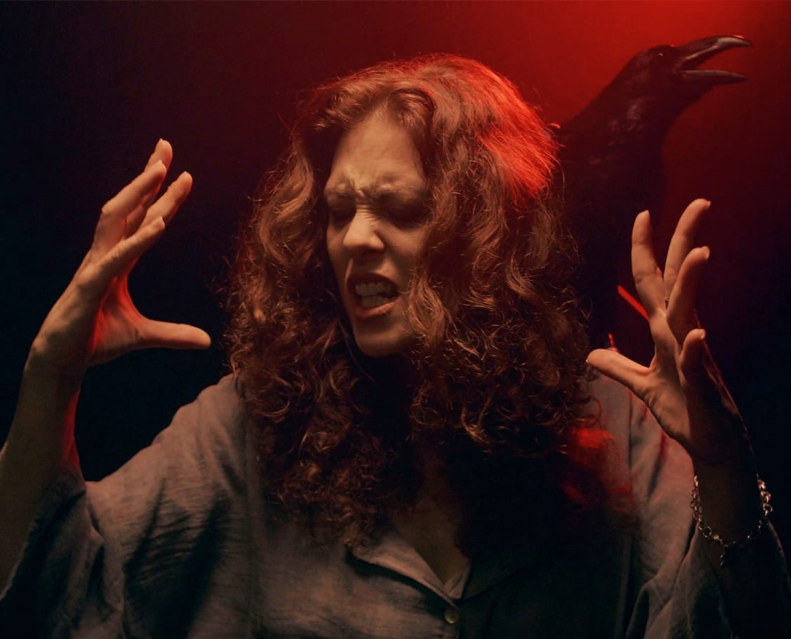 Mujer con cabello rizado y expresión de angustia, gesticulando con las manos, con fondo oscuro y figura de un cuervo en la pared detrás. Interpretando un videoclip de Metal moderno, groove metal, metalcore y deathcore en Madrid.