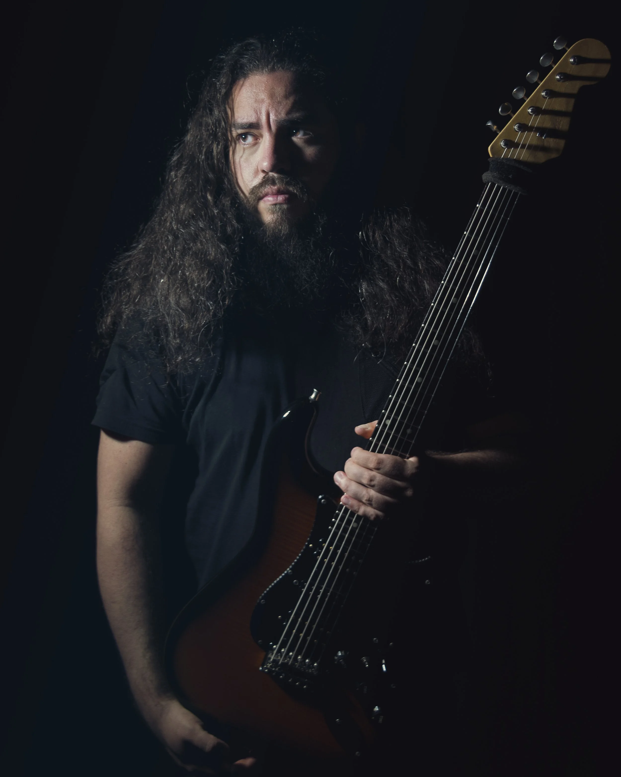 Joven guitarrista fundador de la banda de Metal moderno, groove metal, metalcore y deathcore Rabia Perez Gabriel Peñasco con barba y cabello largo sosteniendo una guitarra eléctrica en un fondo oscuro en Madrid.