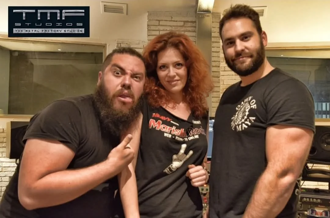 Tres personas en un estudio de grabación, un hombre con barba y camiseta negra en la izquierda, una mujer con cabello rizado y camiseta con estampado en el centro, y un hombre con barba y camiseta negra en la derecha. Detrás hay equipos de sonido y una pared de ladrillos.